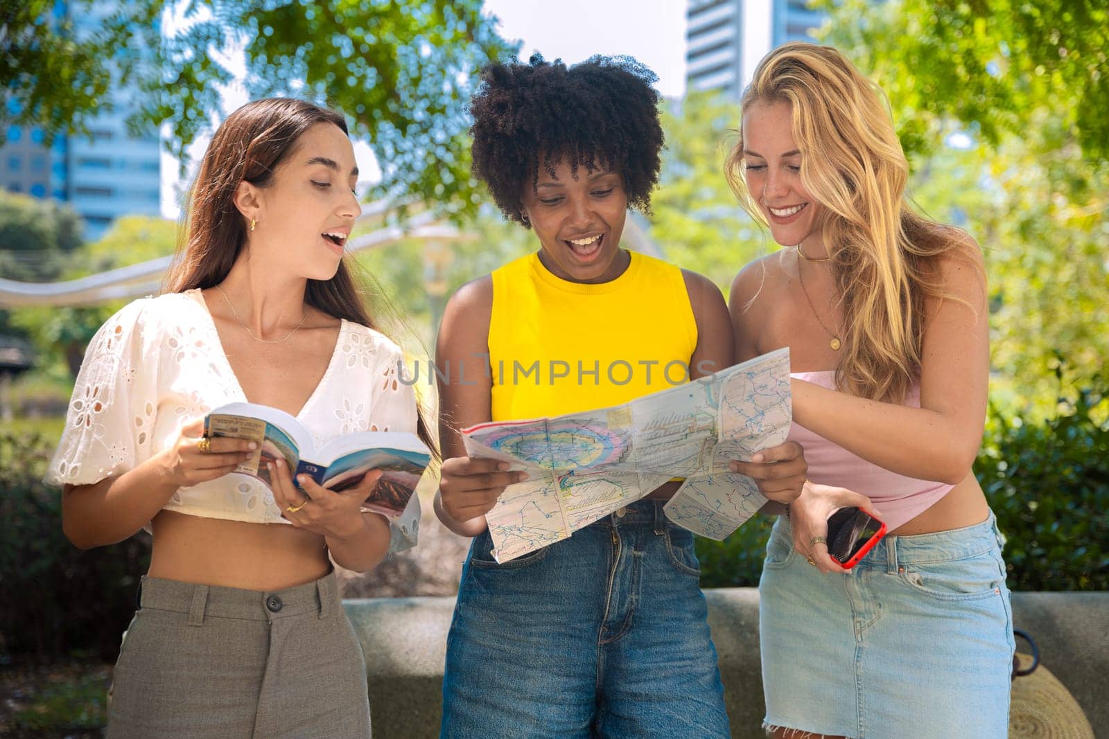 Three happy young women stand outdoors, looking at maps and a travel guide. They smile while planning their trip, enjoying a fun adventure in a sunny urban setting with trees and buildings.