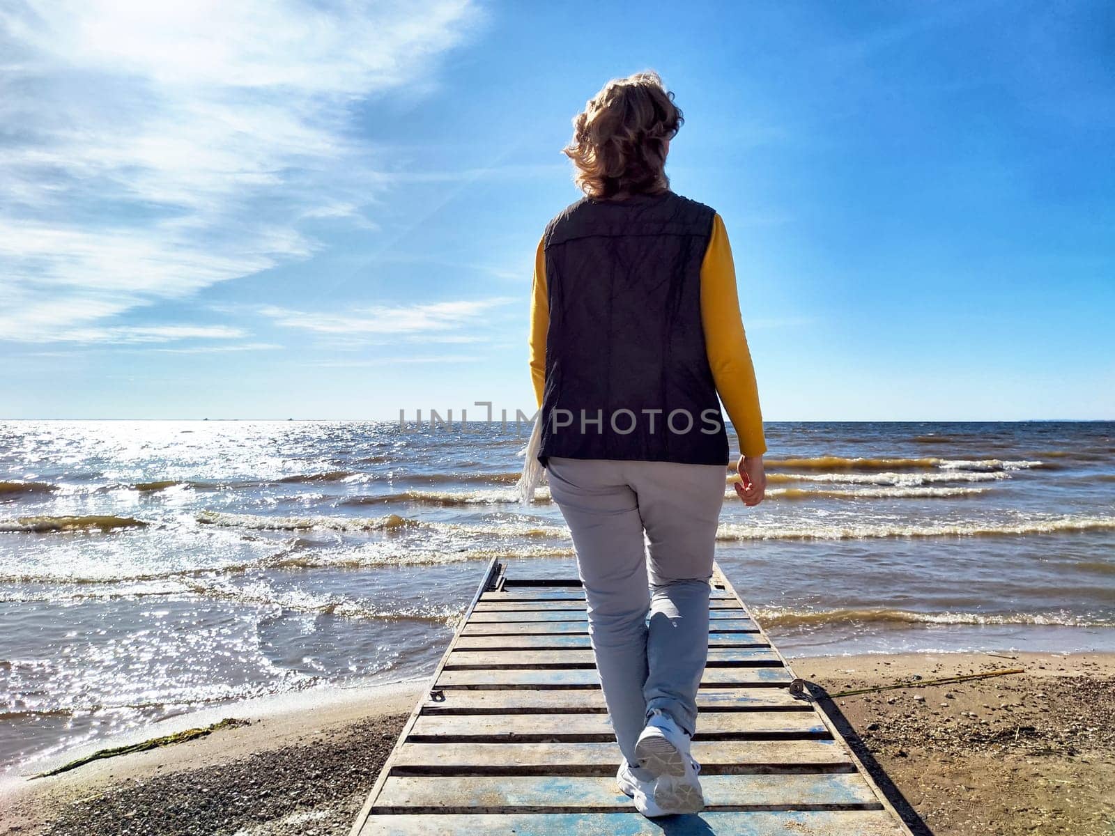 A woman walks along a wooden pier beside the shimmering sea, enjoying a moment of peace and freedom under the bright sky.