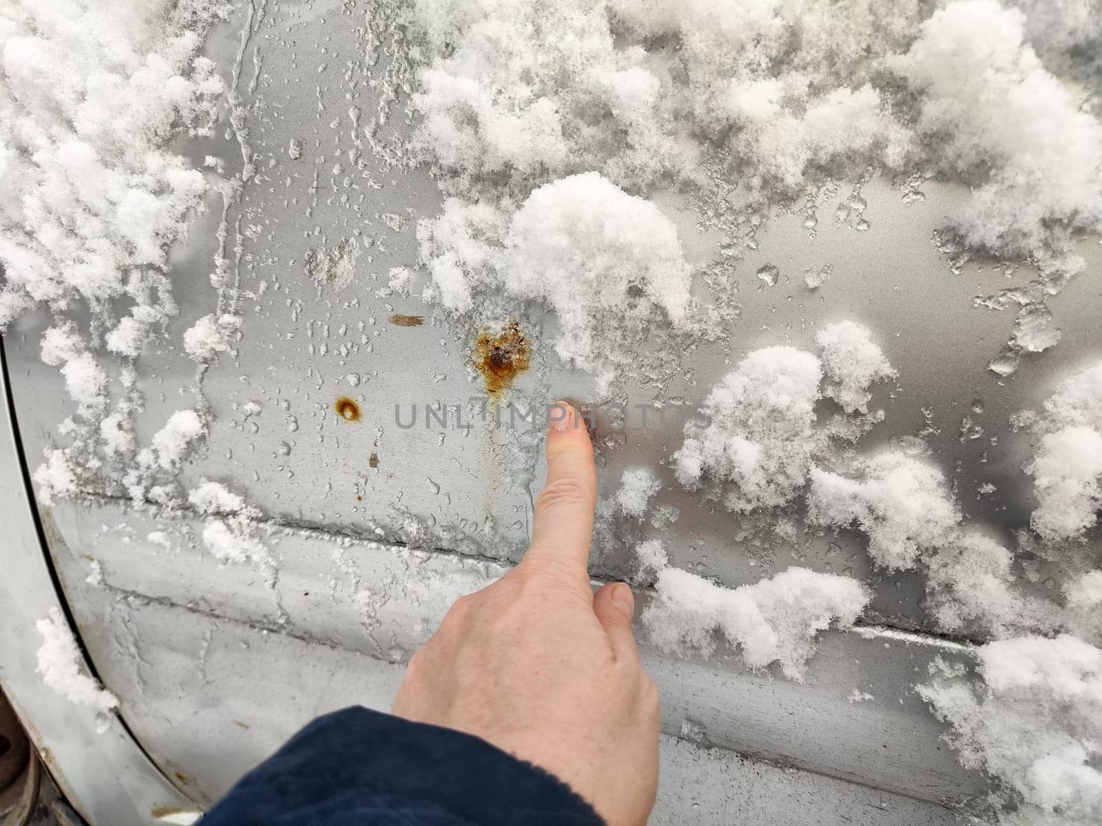 A person points at visible rust spots on a car's exterior covered with snow during cold weather in a neighborhood.