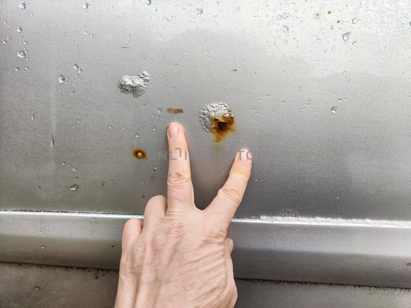 A person examines rust and scratches on silver car parked in a bright outdoor setting, focusing on the damaged area