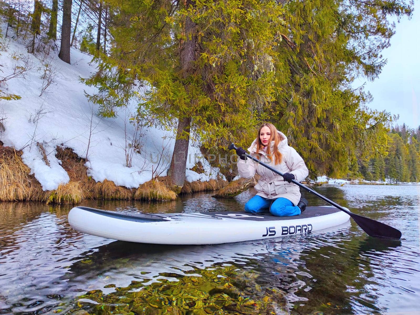 Kirov, Russia - March 14, 2024: A woman enjoys paddleboarding on a calm water body, framed by snow-laden trees in a tranquil winter setting.