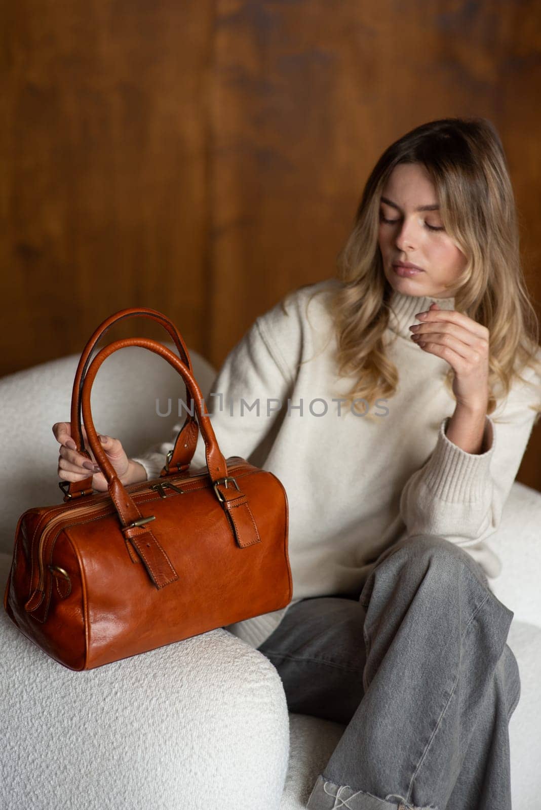 beautiful girl in gray pants and beige sweater posing with a small brown leather bag.