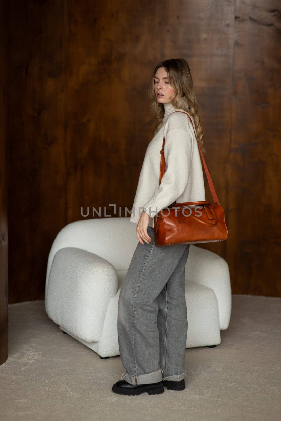 beautiful girl in gray pants and beige sweater posing with a small brown leather bag.