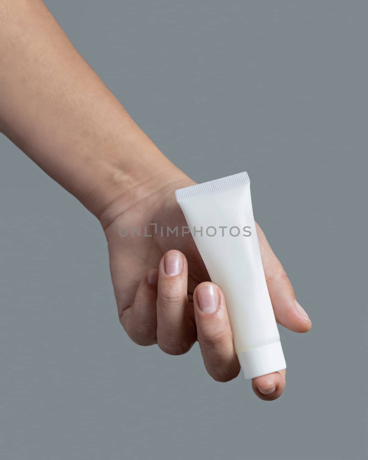 Female hand holding white squeeze cream tube against grey close up, cosmetic package mockup, copy space. Skincare beauty product with minimal composition