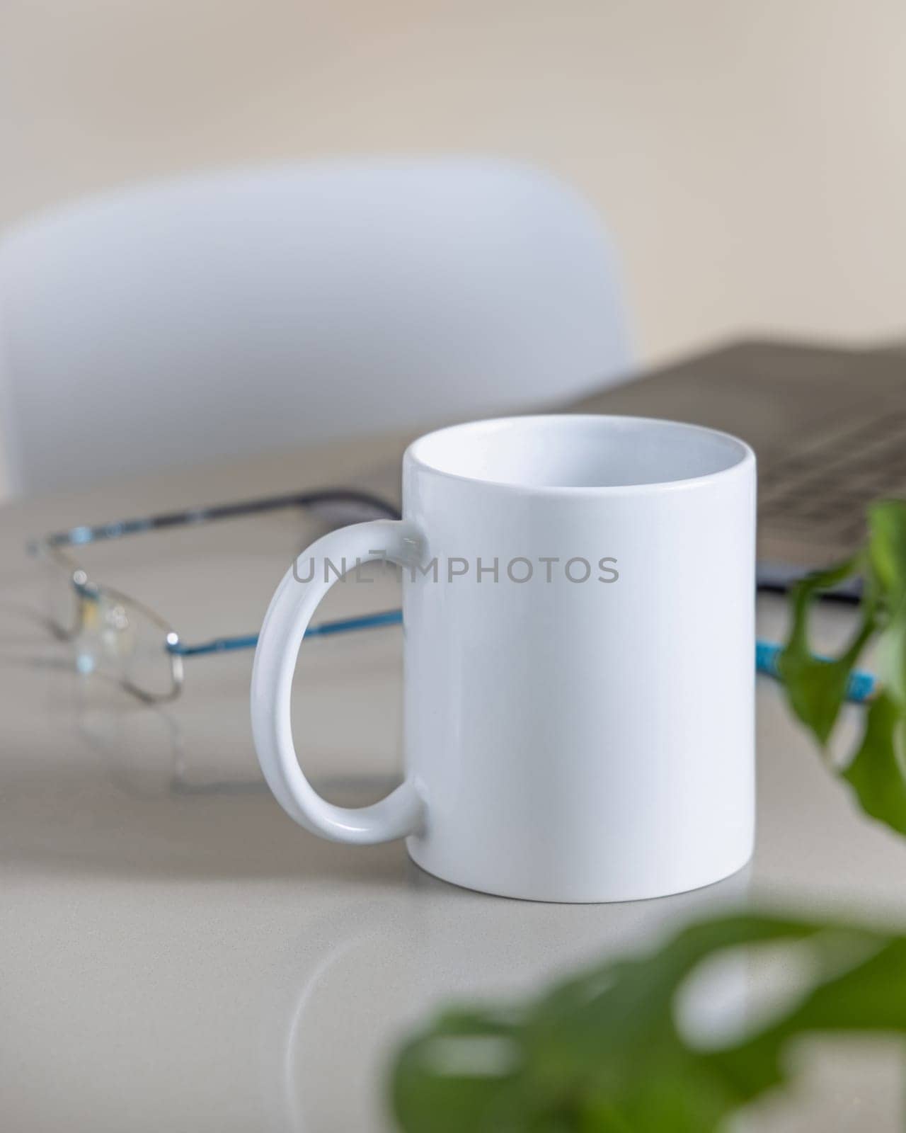 Closeup of 11 oz white blank coffee mug on grey office table against lopened laptop, glasses and green plant, mockup. Template for design with copy space