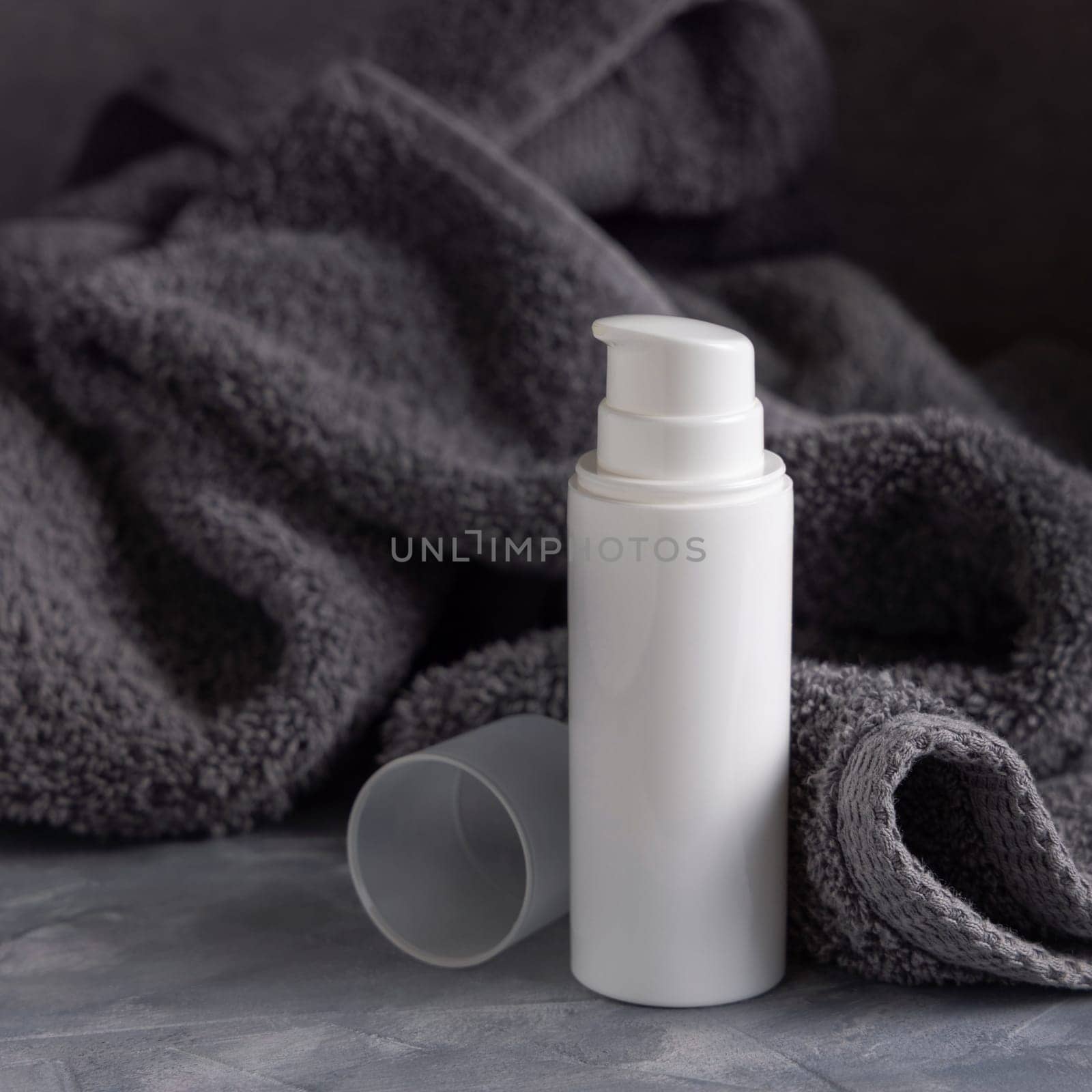 White blank one pump cream bottle near dark grey towel in bathroom closeup, brand packaging mockup, copy space. Natural handmade cosmetic product for facecare and spa procedures