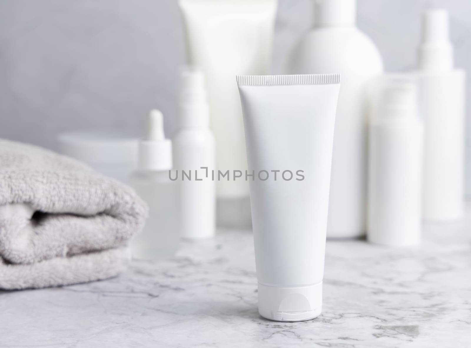 White cream squeeze tube on white marble table against cosmetic bottles and grey towel in bathroom, close up, packaging mockup. Natural product for daily face care, spa procedures