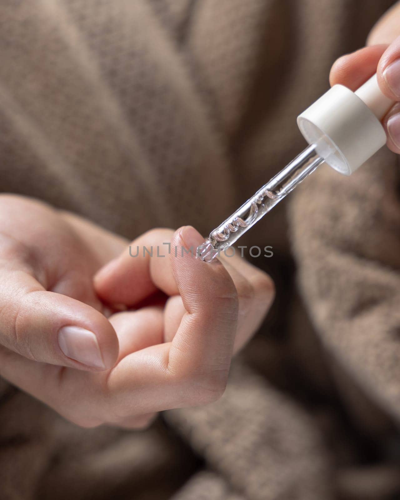 Close up of woman in brown bathrobe dropping cuticle oil on a fingernail and holding a pipette on other hand. Healthy hand skin and nails, natural manicure