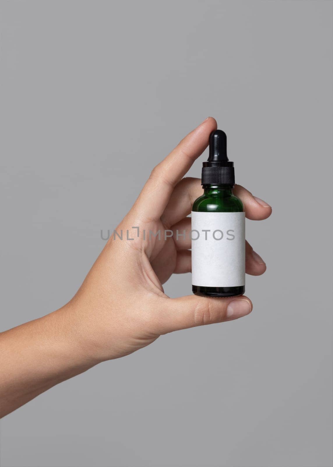 Female hand holding green glass dropper bottle with blank label against grey close up, cosmetic packaging mockup, copy space. Skincare beauty product - serum or essential oil