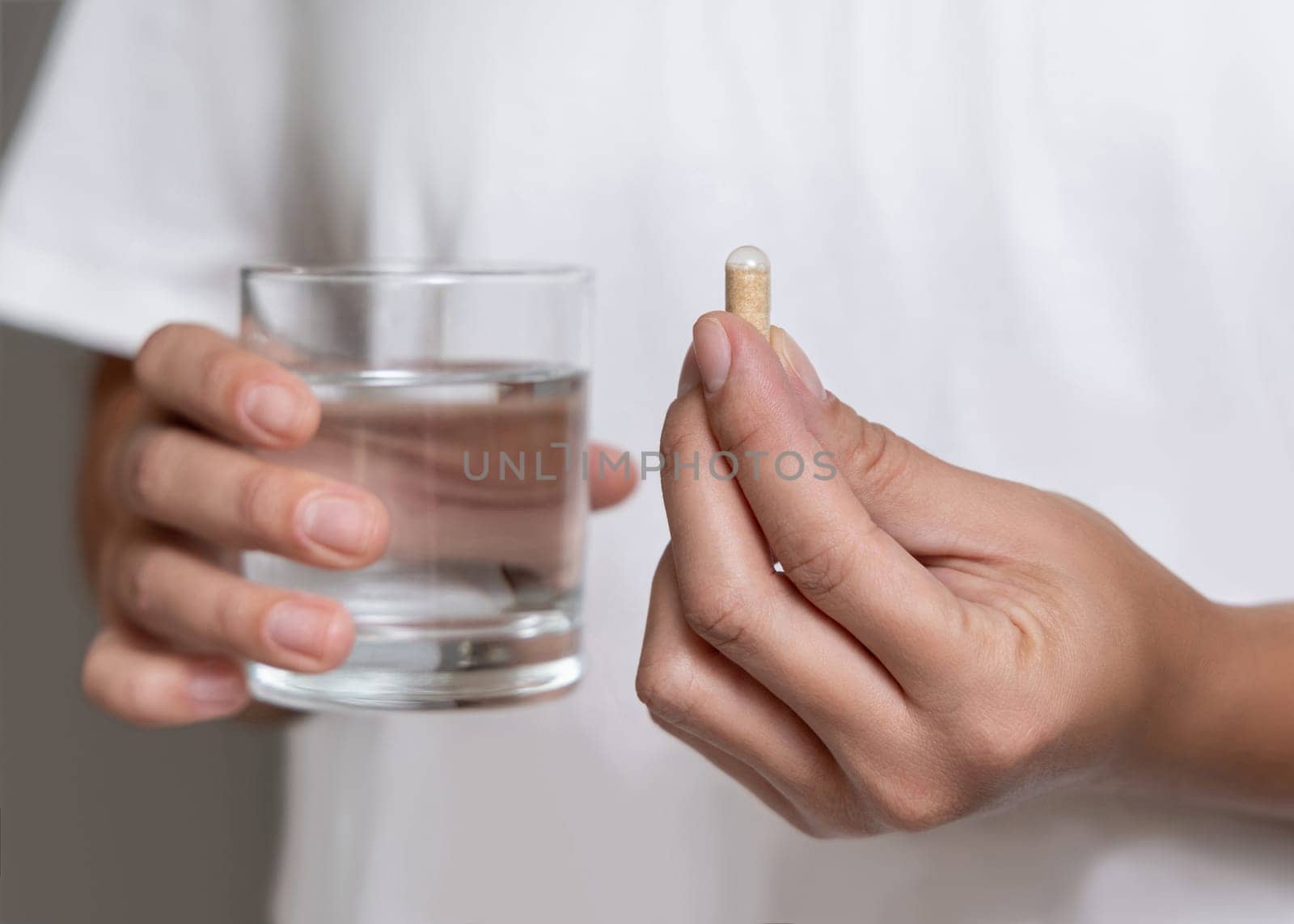 Woman in white t-shirt holding beige capsule in fingers and water glass in hand closeup. Female or male taking dietary supplement, daily vitamins. Healthcare concept
