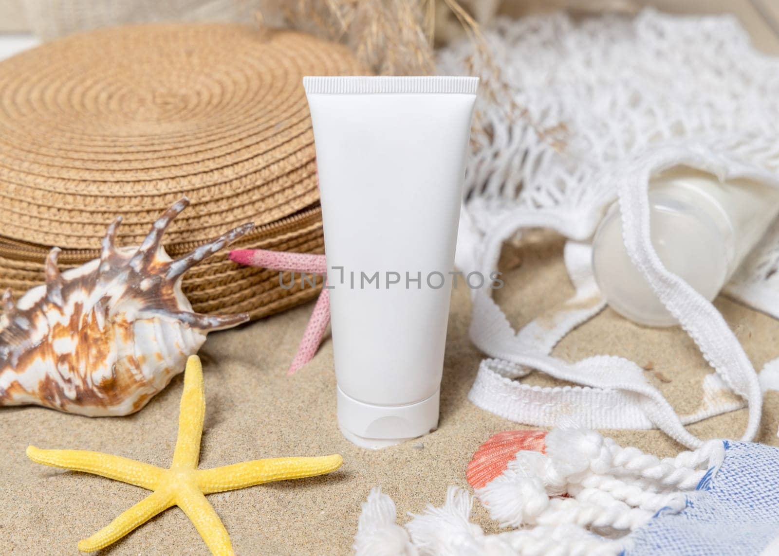 White blank cream tube, bag, towel, starfishes and seashells on a sandy beach, closeup. Sunscreen cosmetics mockup. Summer vacation with spf uv-protected sunbathing