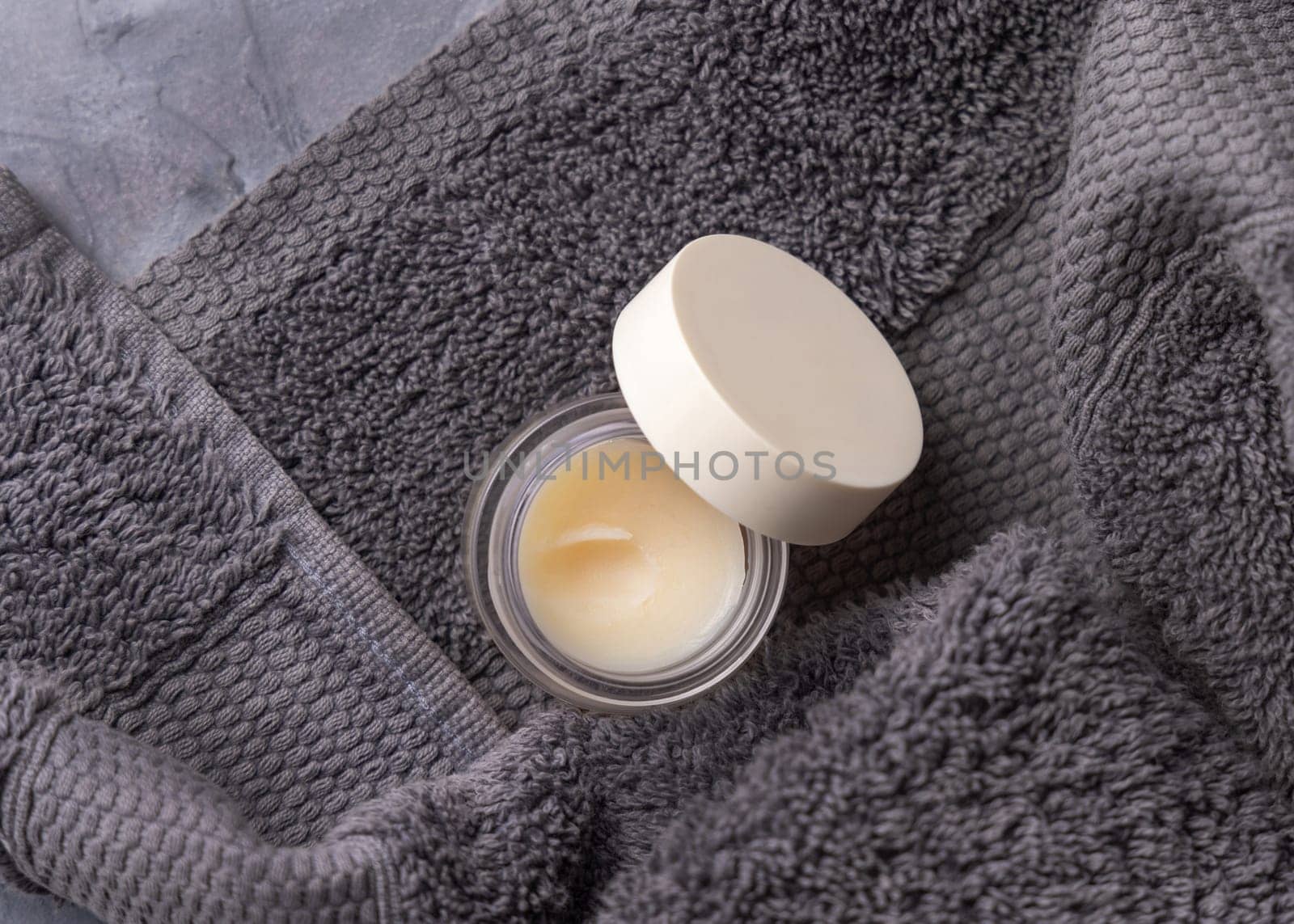 Opened lip balm jar with blank lid near dark grey folded towel top view, cosmetic packaging mockup, copy space. Lipcare beauty product for mens everyday personal care