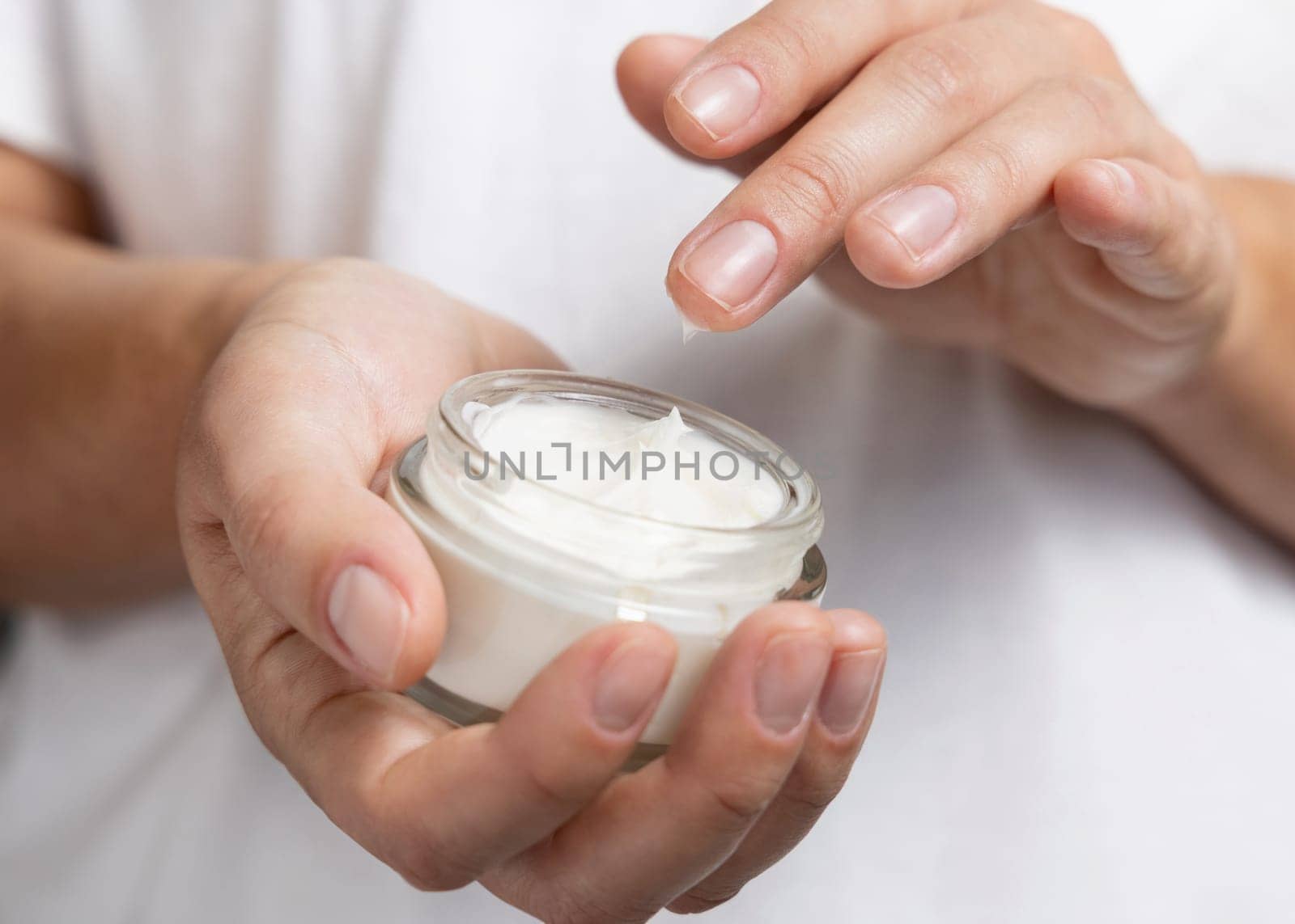 Close up of woman touching cream with finger and holding a cosmetic jar in other hand with natural nanicure agains white shirt. Concept of organic cosmetics for body care