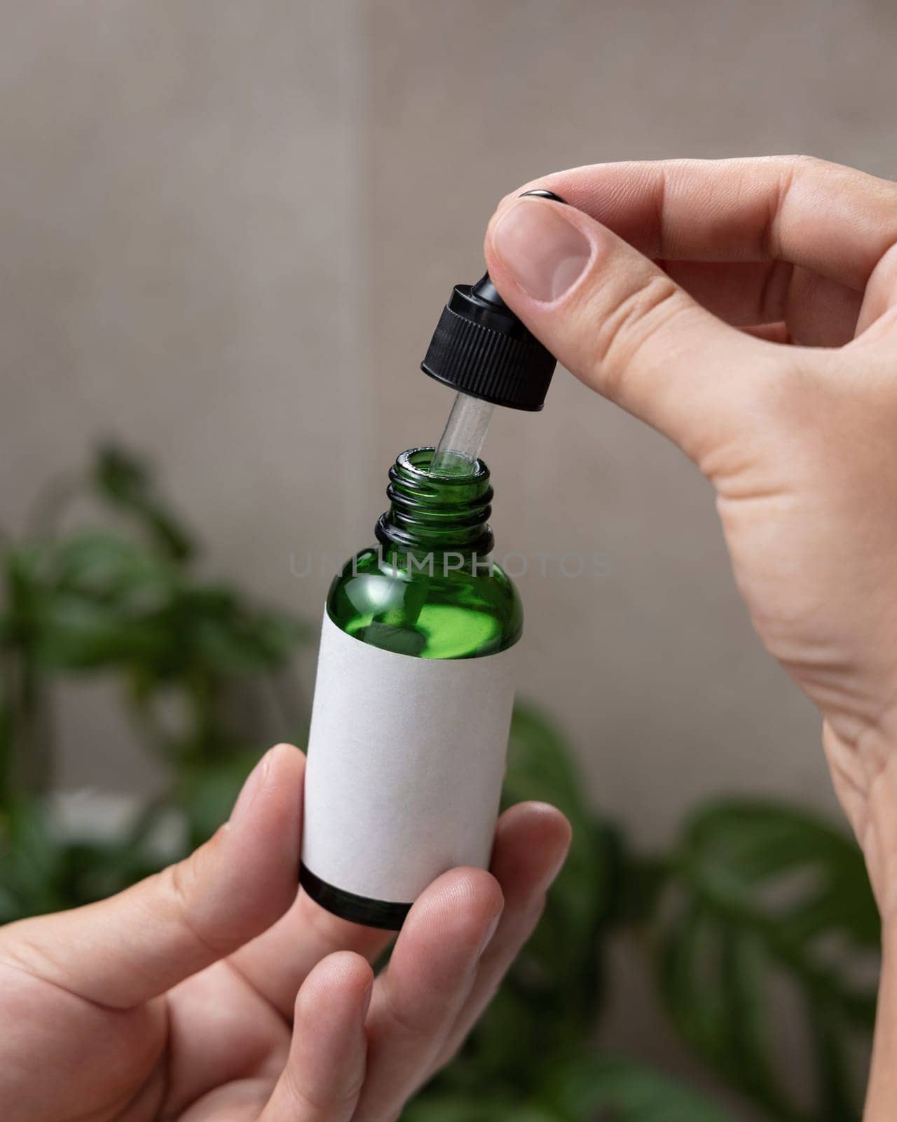 Female hands with natural manicure opening green dropper bottle with blank label, close up against green plant. Mockup for cosmetic bottle with pipette, serum or essence. Beauty spa spa procedures