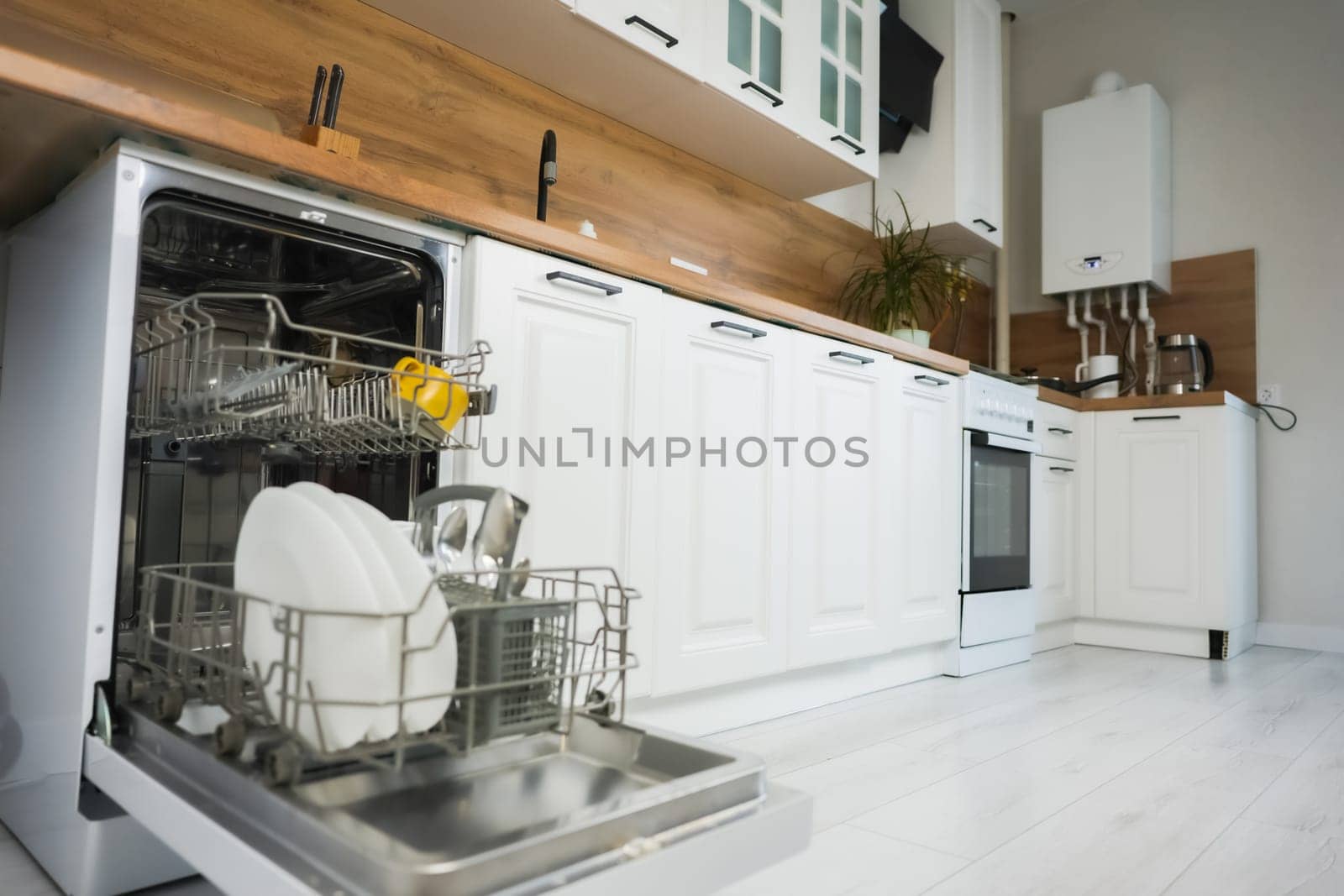 Open dishwasher with dish drawer pulled out close-up on the left, with clean dishes and cutlery in the background of a white and wooden kitchen in a slight out of focus. Comfort concept, dishwashing. High quality photo