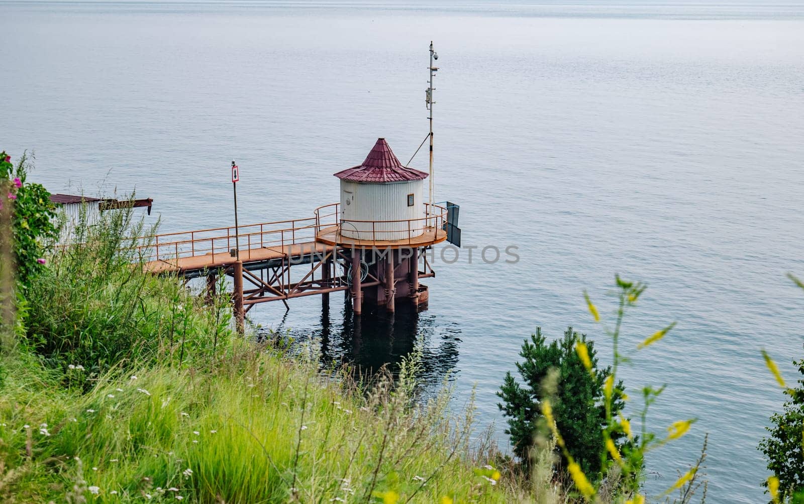 Small white water intake building standing on stilts in a calm russian lake, connected to the shore by a metal pier and surrounded by lush vegetation on an overcast day