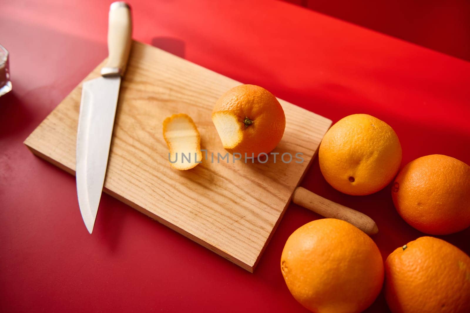 Fresh whole and sliced oranges displayed on a wooden cutting board with a kitchen knife, placed atop a red surface, showcasing vibrant colors and a kitchen preparation scene.