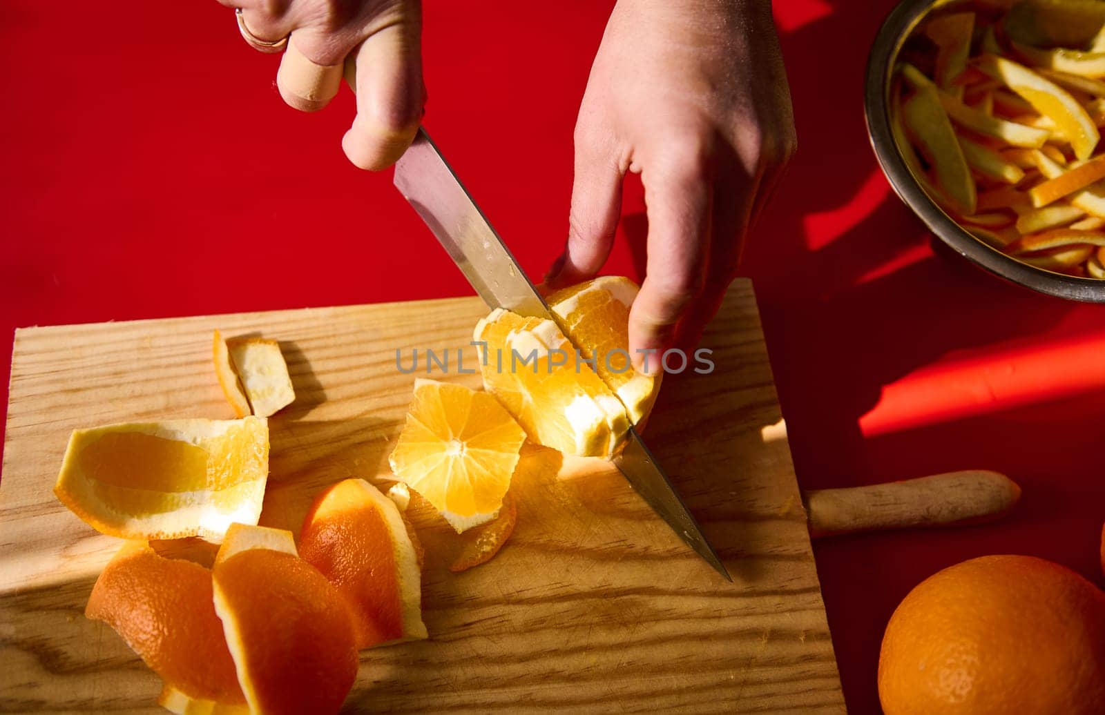 A close-up view of hands slicing a peeled orange on a wooden cutting board, emphasizing fresh fruits and the culinary process against a vivid red background.
