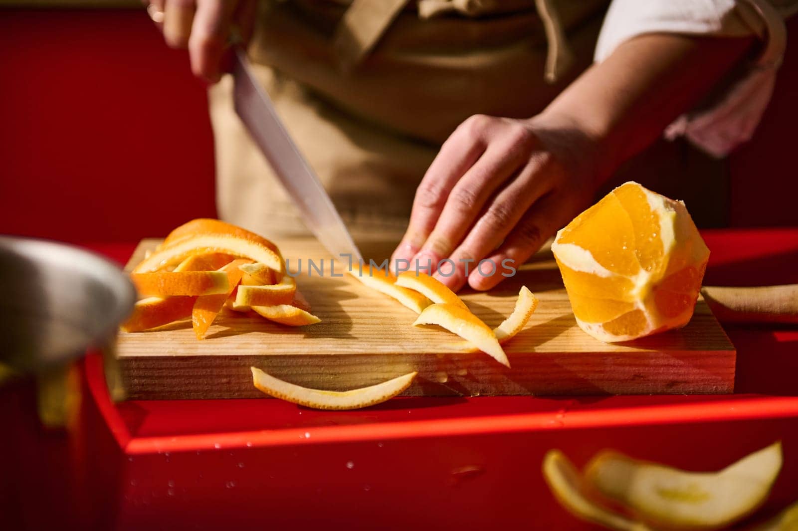 A person carefully slices an orange peel on a wooden cutting board, capturing culinary preparation.