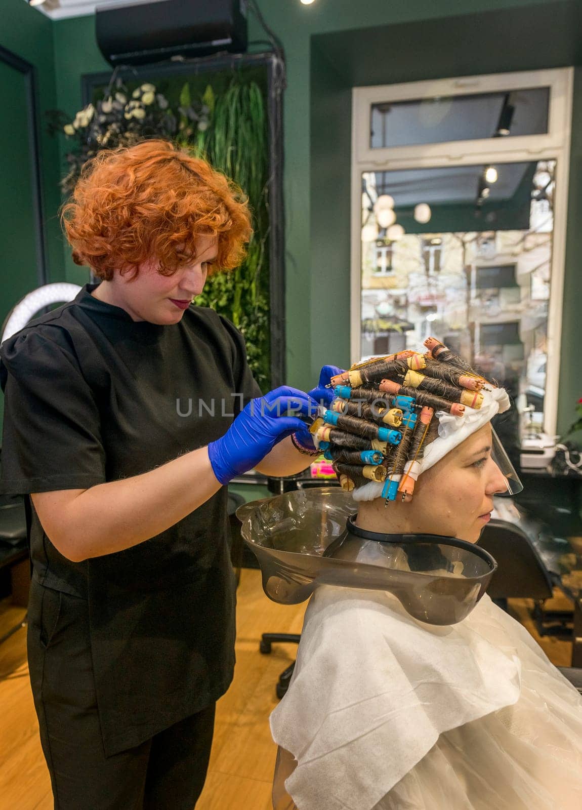 A young girl is getting her hair curled with curlers by a hairdresser. The client is sitting in a chair and her hair is curled with curlers. The hairdresser is curling the curlers