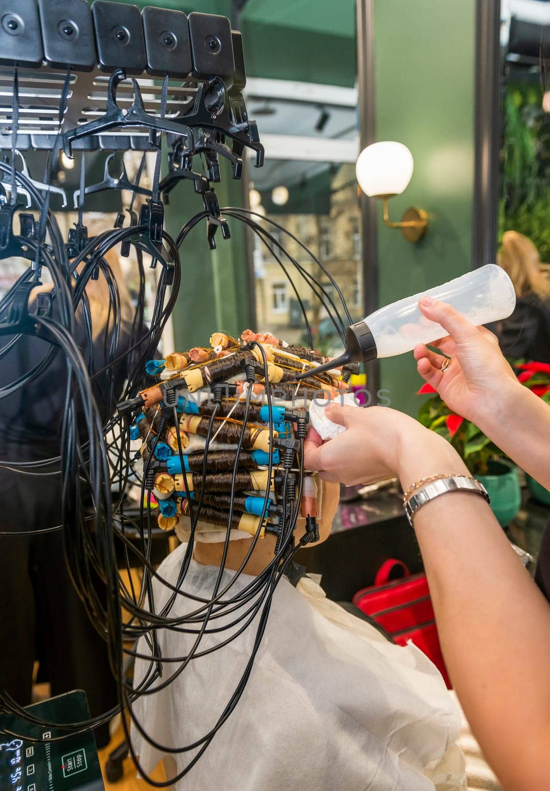 modern process of hair treatment and curling. client sits in chair with curlers on her head. machine treats and makes hair curly and wavy. master applies liquid to hair, close-up