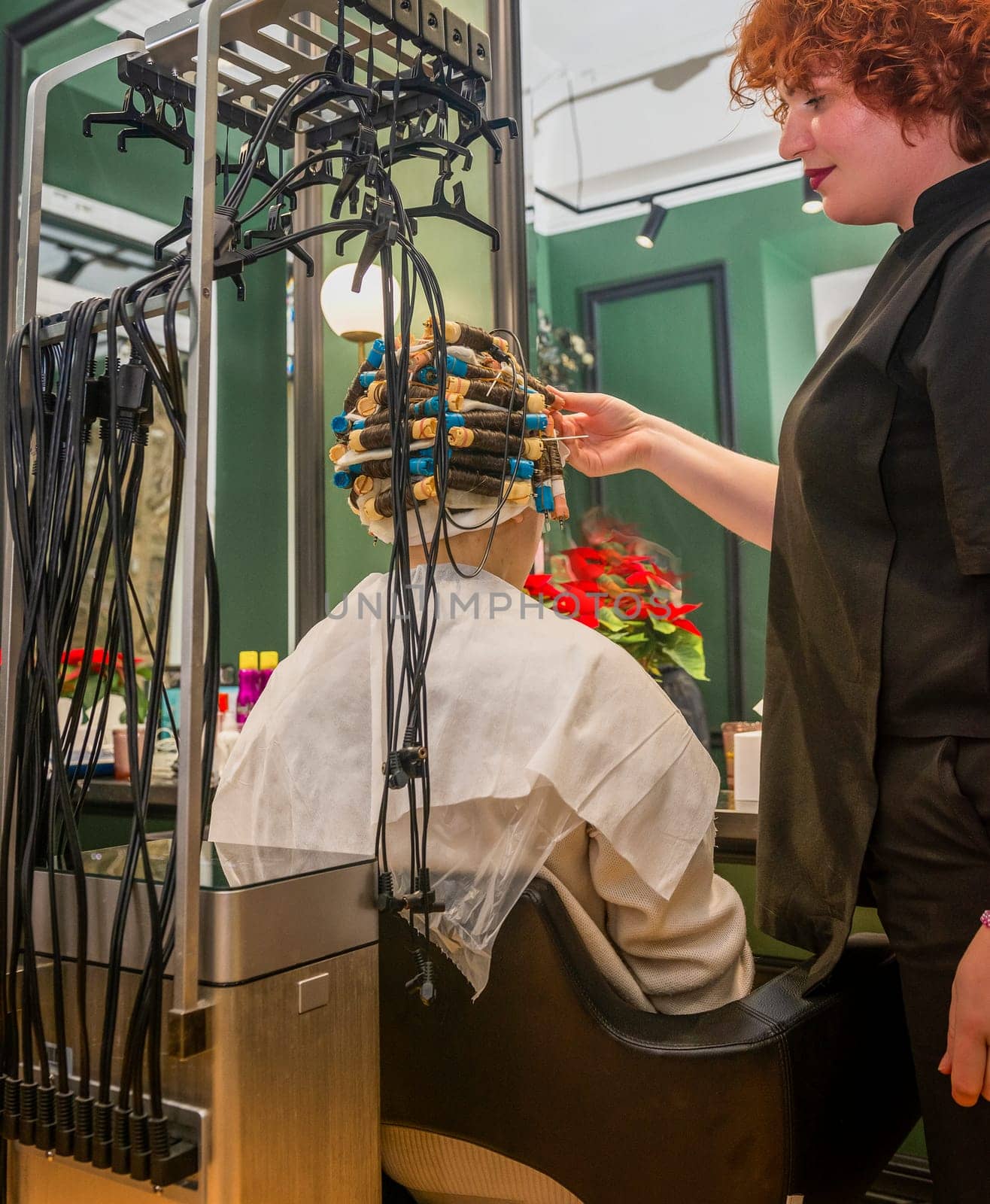 the client is sitting in a chair with curlers on her head the device treats and makes hair curly and wavy. the master adjusts the curlers the device is connected