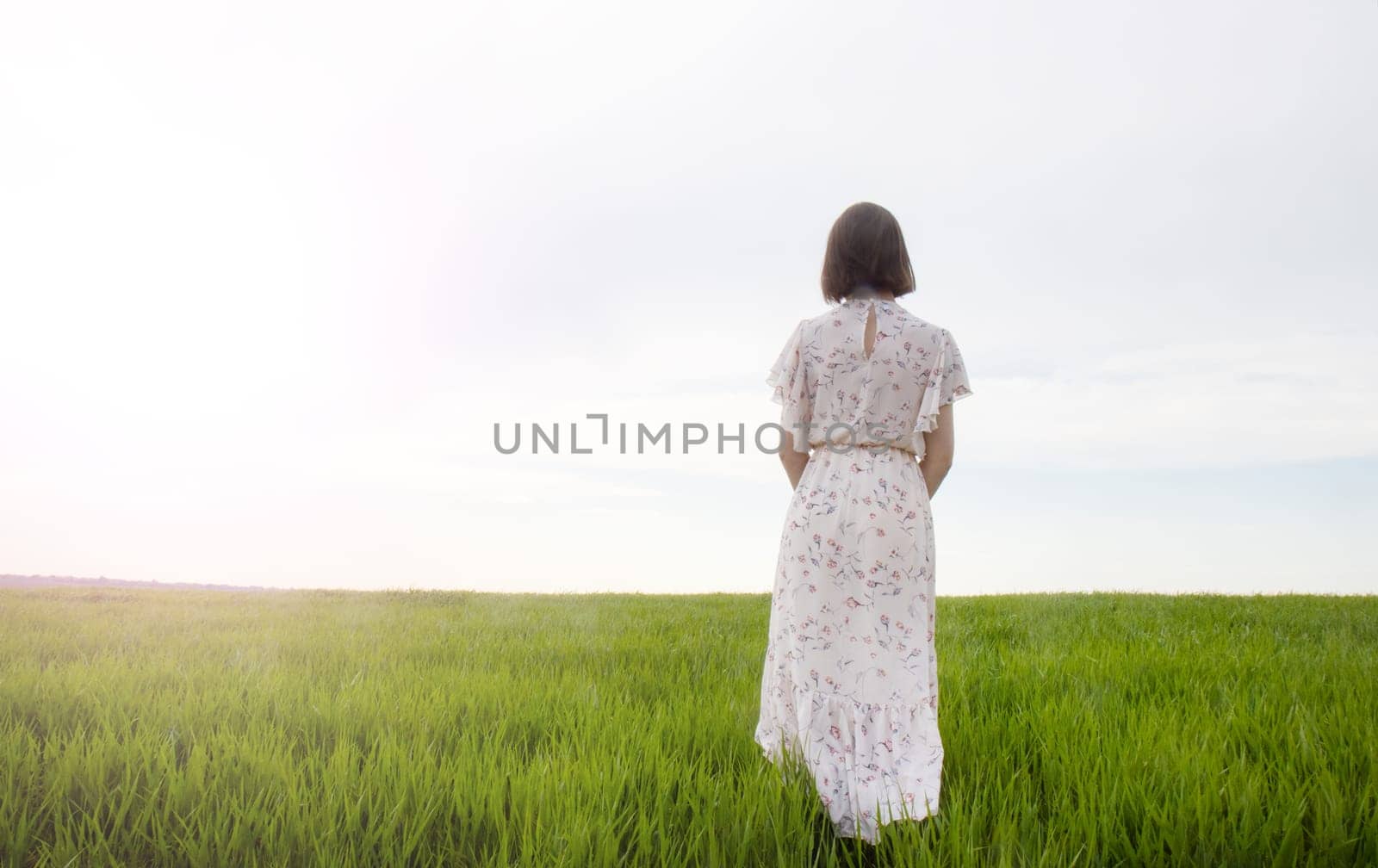 A woman in a floral dress stands gracefully in a green field, gazing at the bright sky and exuding tranquility