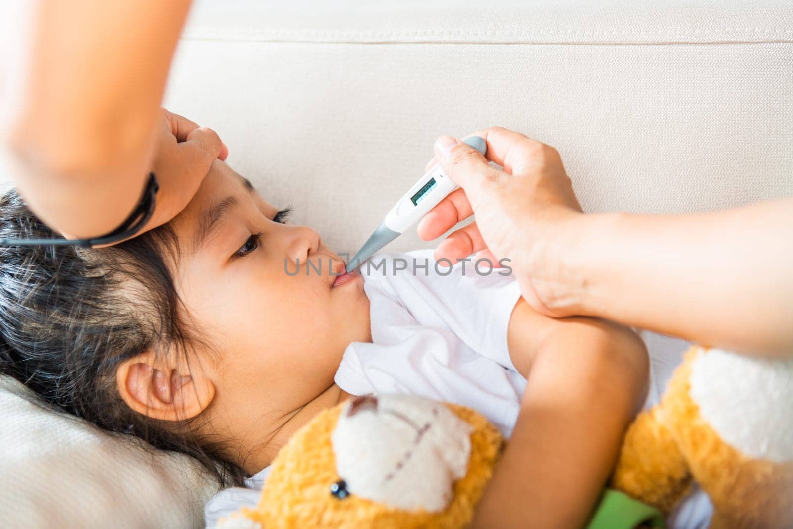 Sick kid. Mother checking temperature of her sick daughter with thermometer in mouth, child laying in bed taking measuring her temperature for fever and illness