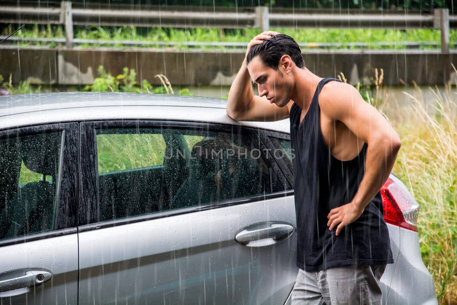 A man standing next to a car in the rain, sad because his vehicle broke down
