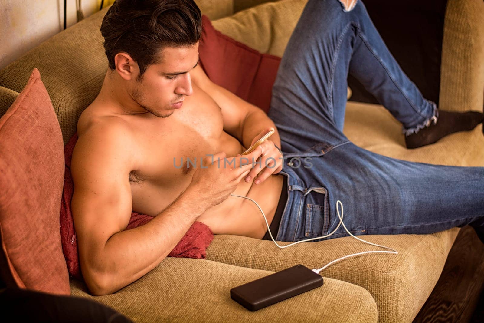 A shirtless man sitting on a couch using a cell phone, recharging it with a power bank device