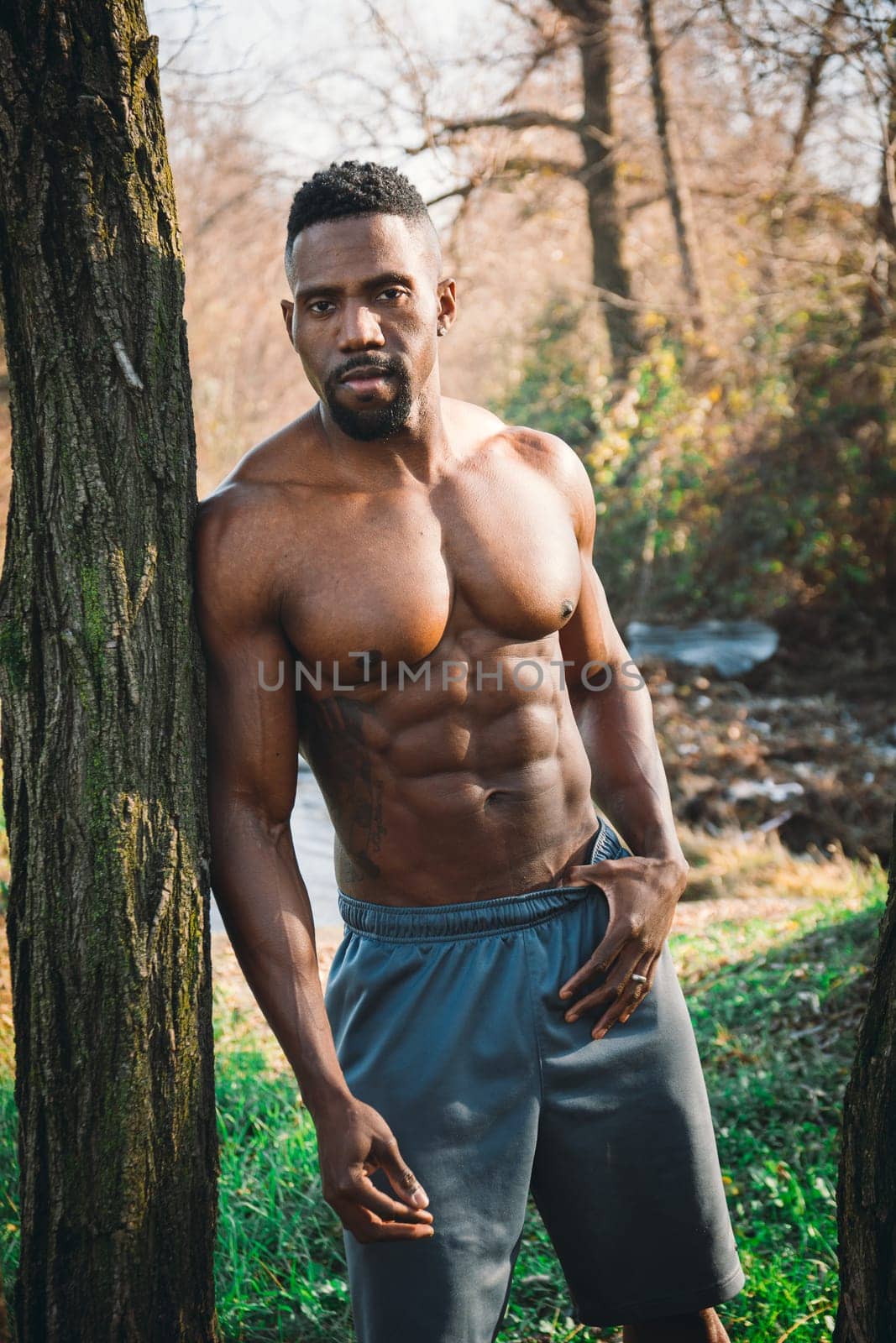 Photo of a shirtless man standing next to a tree in a natural setting by artofphoto