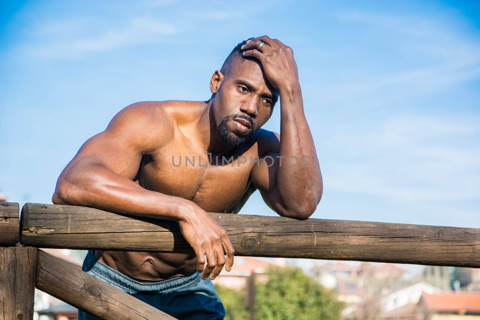 A shirtless muscular african american man leaning on a wooden fence in a city park in a sunny day