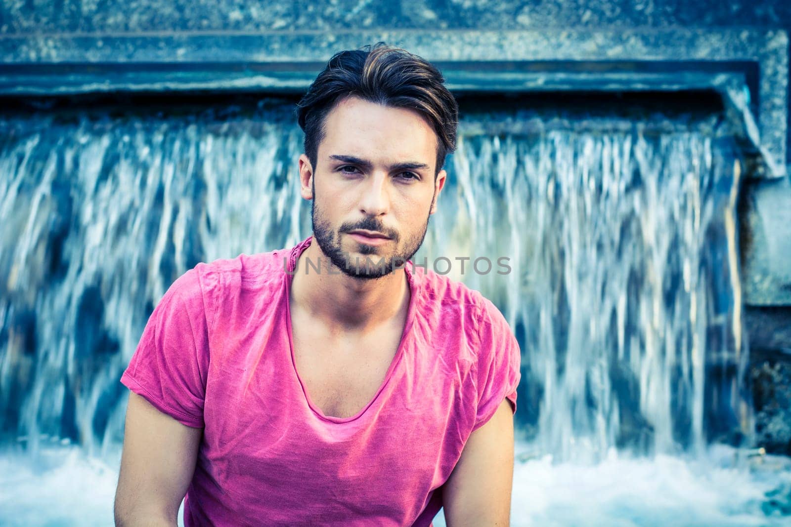 A man in a pink shirt sitting in front of a fountain. Photo of a man in a pink shirt enjoying the serene beauty of a waterfall shaped fountain