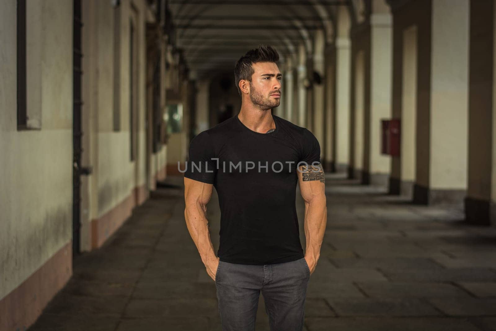 A man in a black shirt standing in a cloister. Photo of a handsome muscular man standing in a colonnade, outdoor in a city, wearing a black shirt