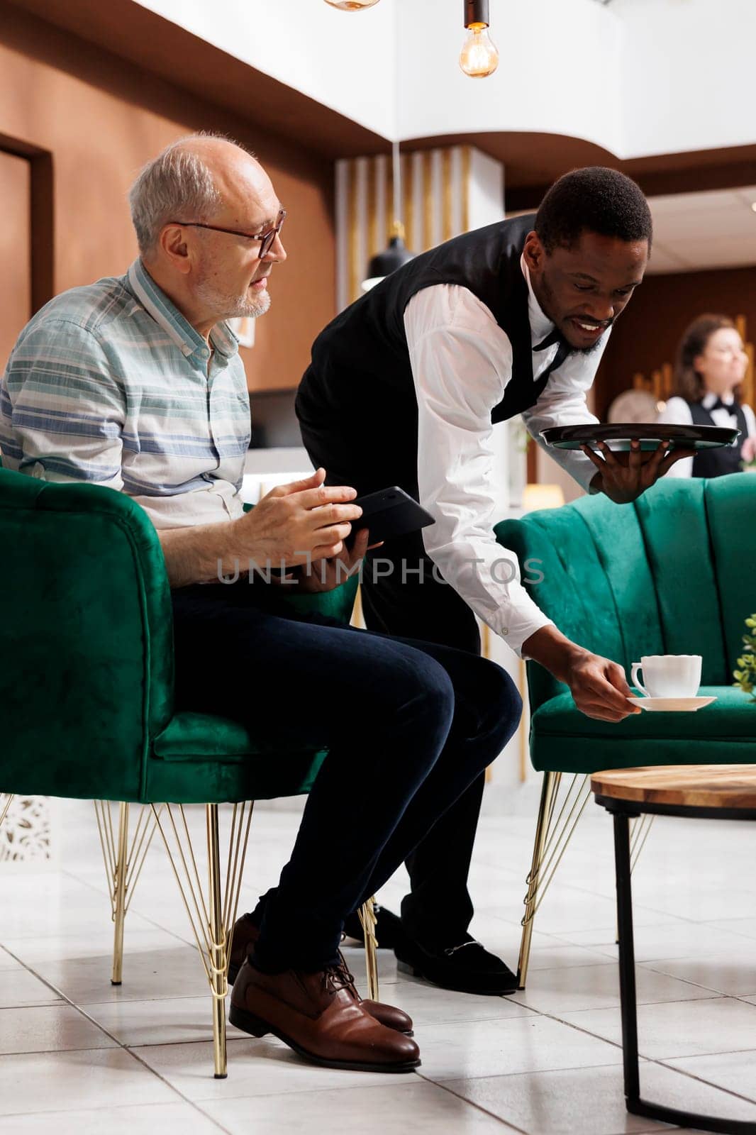 In lounge area, nice waiter serving cup of tea to elderly male tourist seated on couch with tablet. Retired senior man sitting on cozy sofa and being attended by african american staff in hotel lobby.