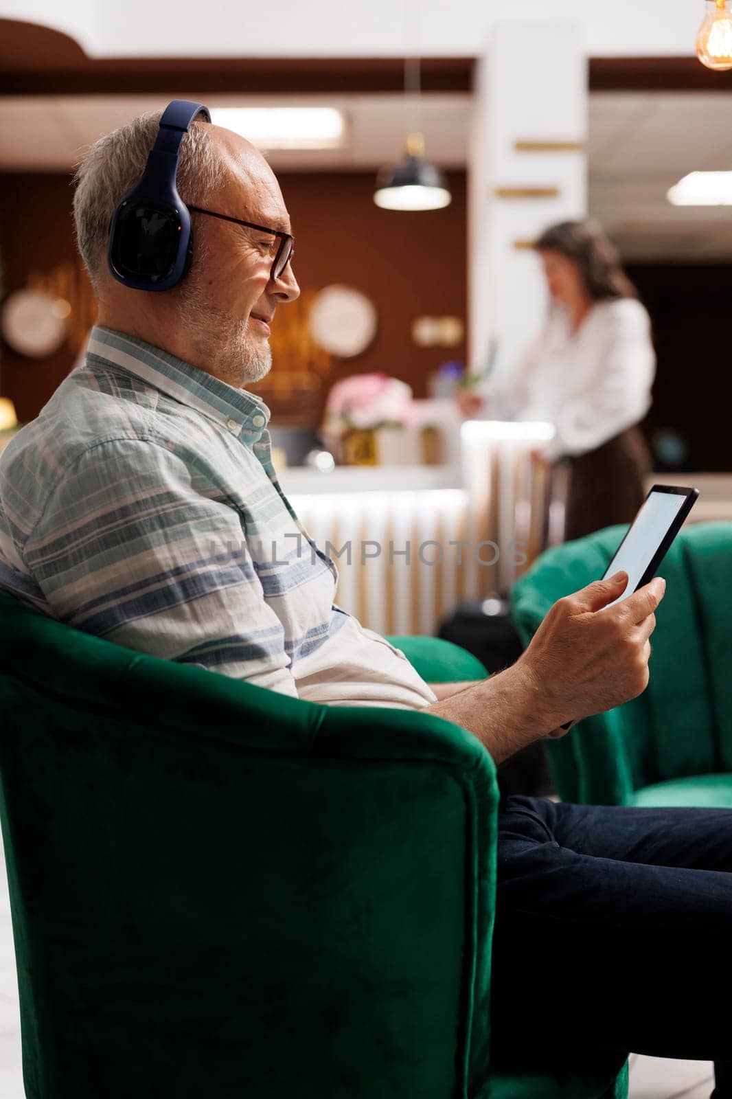 Elderly caucasian man wears headphones and sits on hotel lobby sofa grasping tablet. Senior male guest having wireless headset waits in lounge area and uses digital gadget to browse the internet.