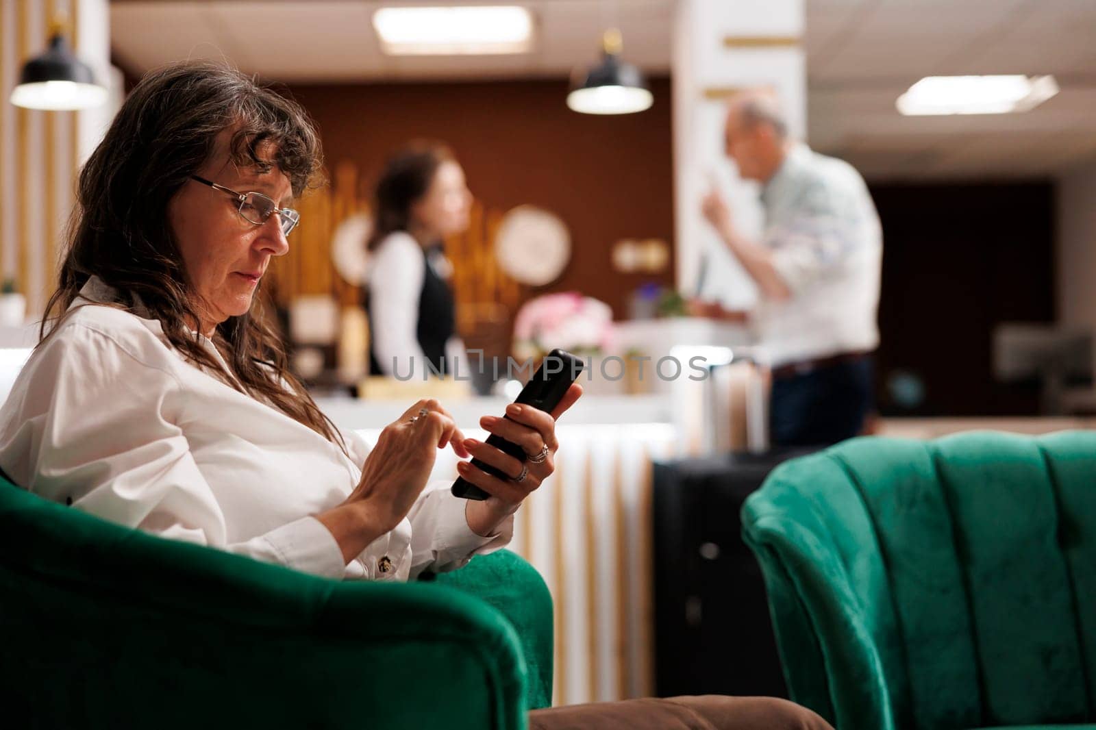 Retired senior woman enjoys online connectivity in hotel lobby, using mobile phone for communication and relaxation. Elderly female traveler relaxing with smartphone in luxury lounge area.