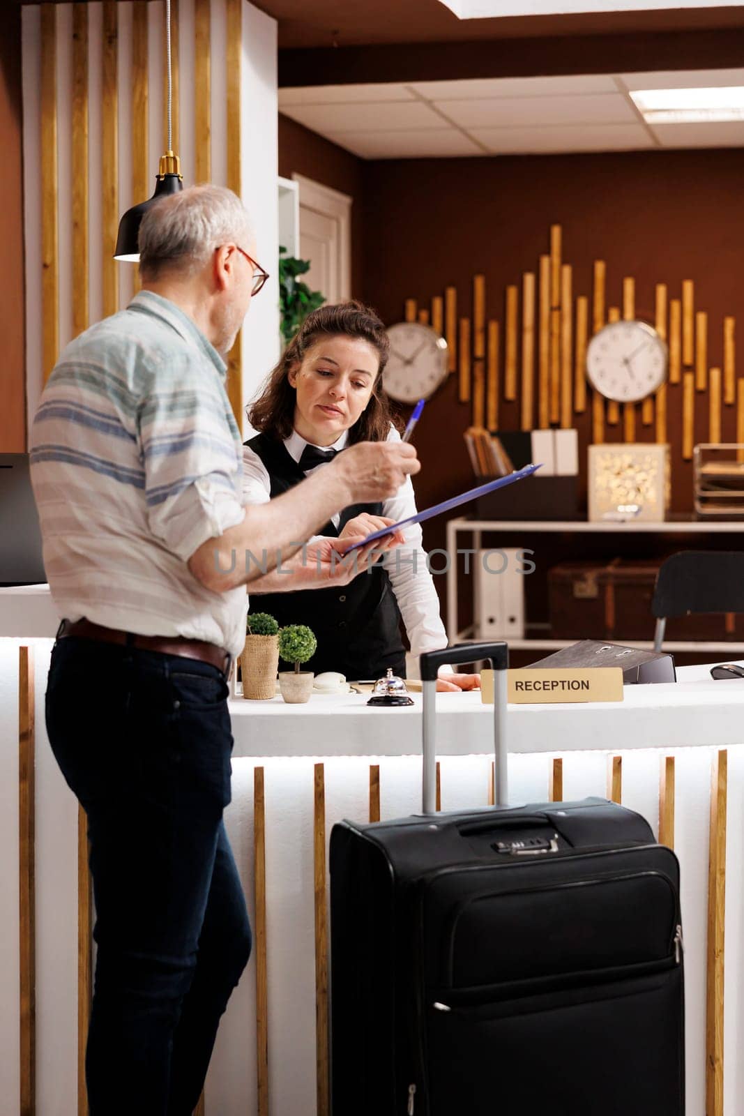 Senior man tourist checks in at the front desk of a hotel, eager for a pleasant holiday. While filling out booking reservation documents, a kind female concierge advises an old man traveller.