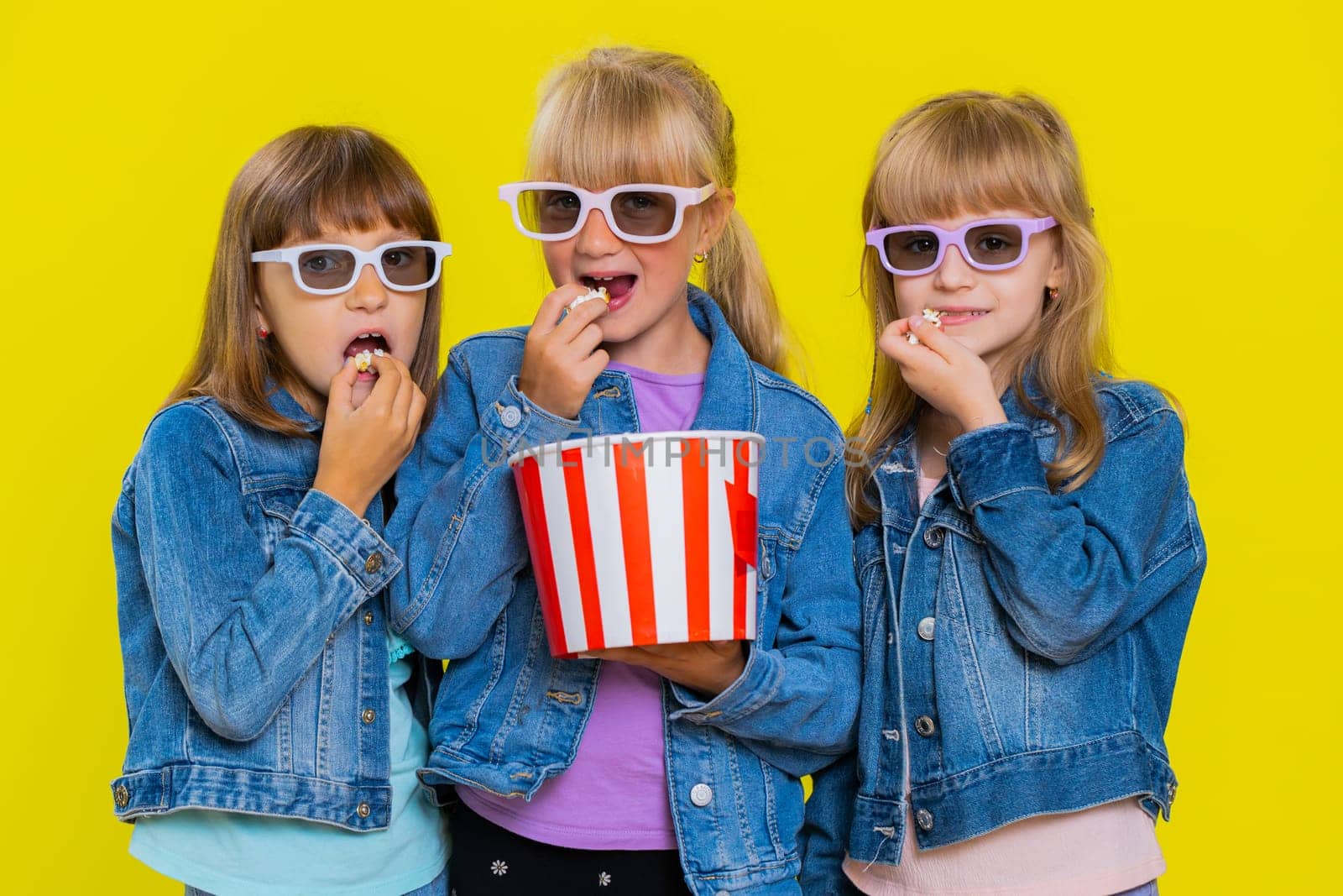 Teenage girls wearing 3D glasses eating popcorn watching interesting tv serial, sport game, film, online social media movie content. Little children sisters. Three siblings kids on yellow background