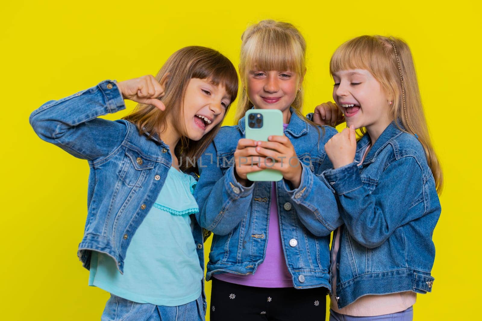 Teenage girls use mobile smartphone typing browsing say Wow yes found out great big win good news celebrate, playing online game. Little children sisters. Three siblings kids on yellow background