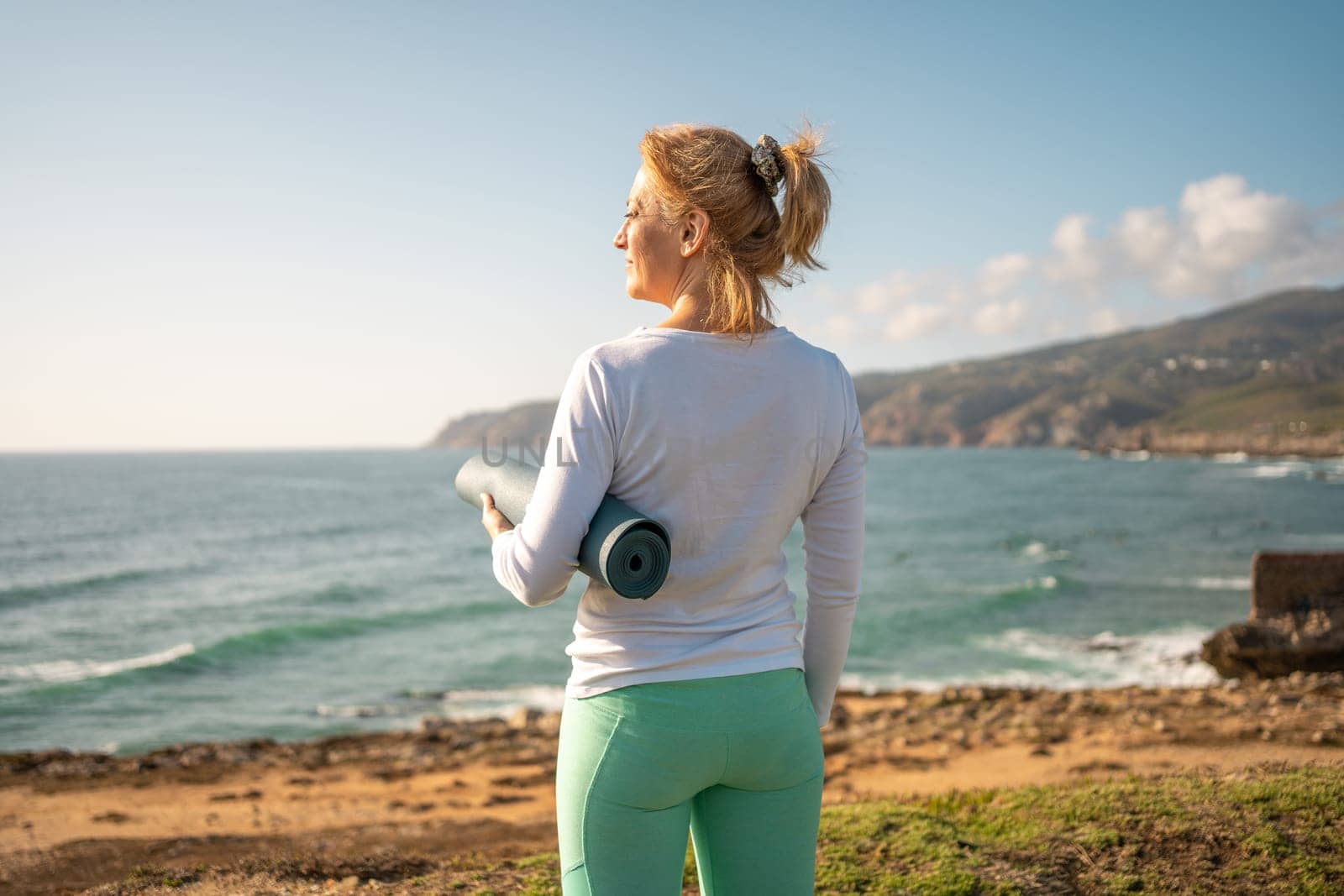 Mature woman with yoga mat standing on coast. Pensioner female looking ocean waves while holding sport mat.Back view fit senior woman wearing sport clothes preparing to do yoga on sea shore