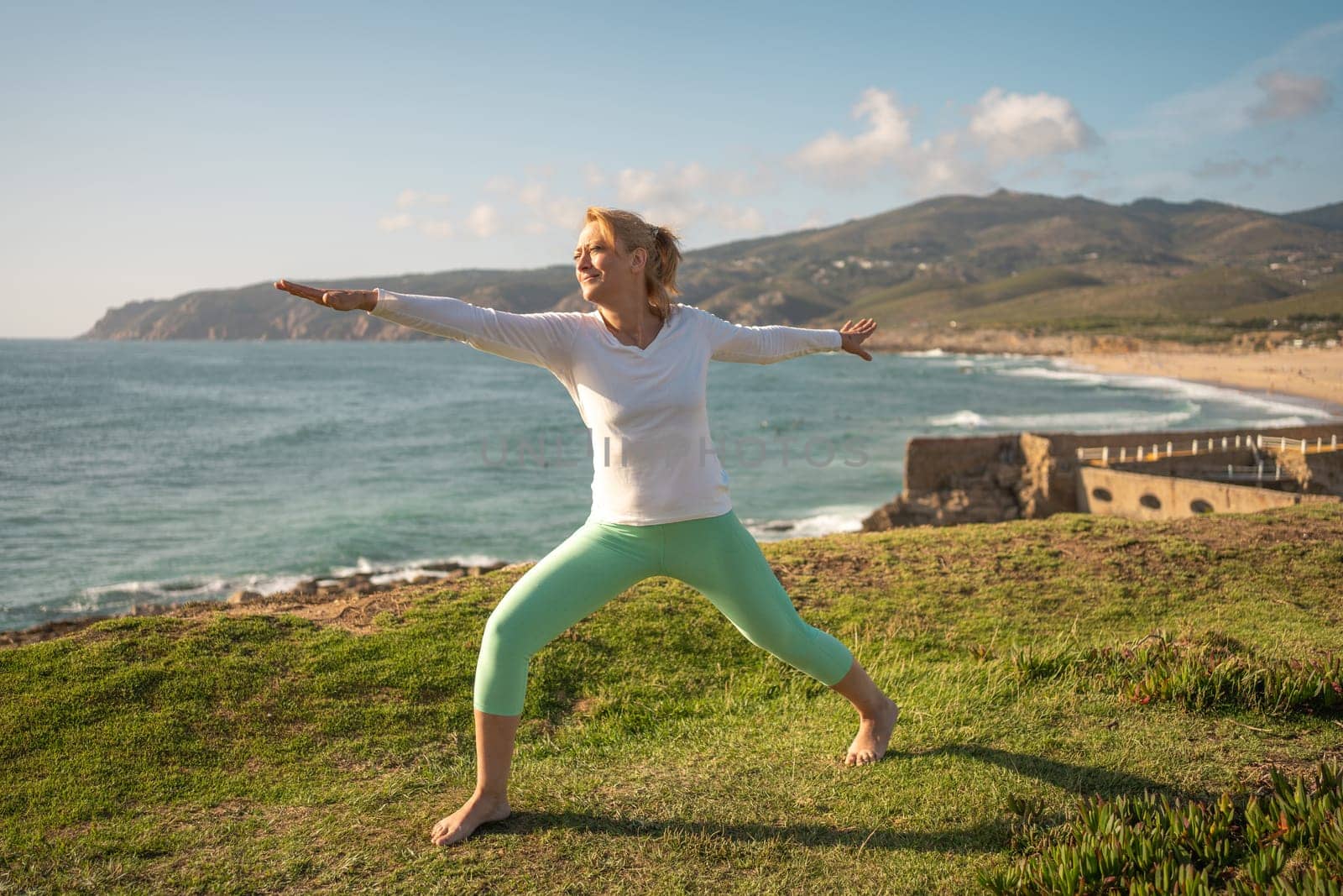 Senior woman practicing yoga on coast. Female standing in warrior position on sea shore with an ancient fortress on background. Mature woman barefoot doing workout near medieval fortification on ocean beach.
