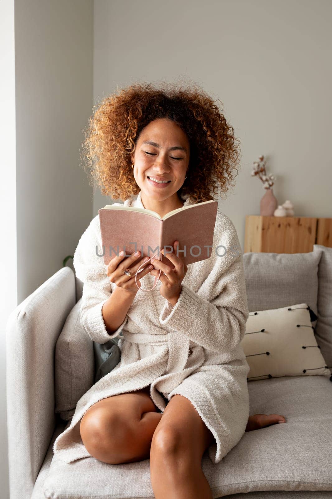 Vertical portrait of happy hispanic woman reading personal journal sitting on the couch at home. by Hoverstock