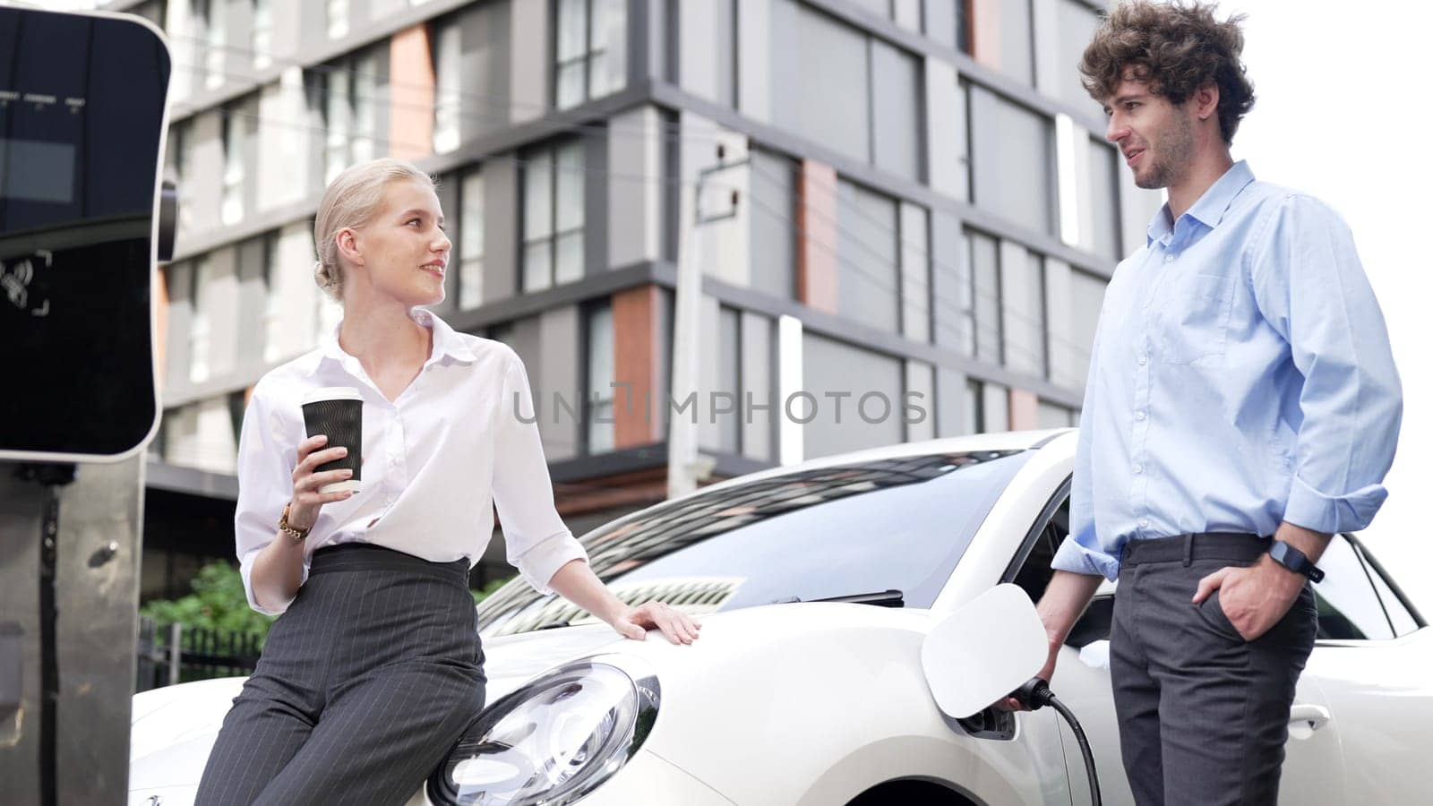 Progressive businessman and businesswoman leaning on electric car connected to charging station before driving around city center. Eco friendly rechargeable car powered by clean energy.