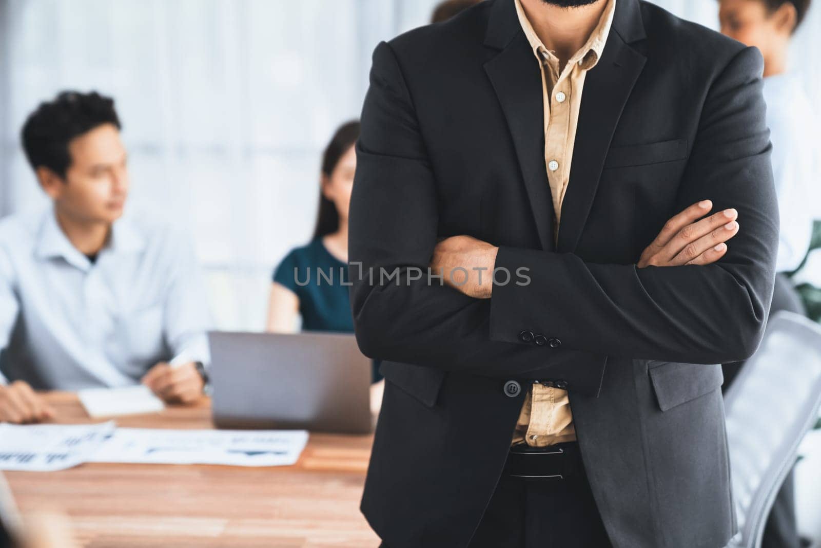 Businessman poses confidently with diverse coworkers in busy meeting room background. Multicultural team works together for business success. Modern businessman portrait. Concord