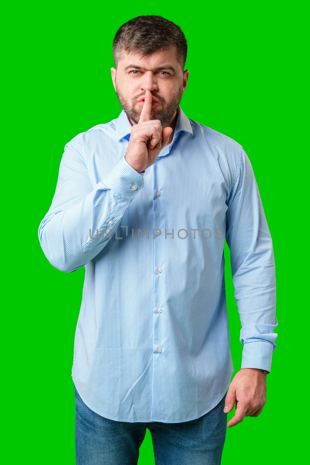 A man in a blue shirt gestures for silence with a finger on his lips against a bright green background in studio