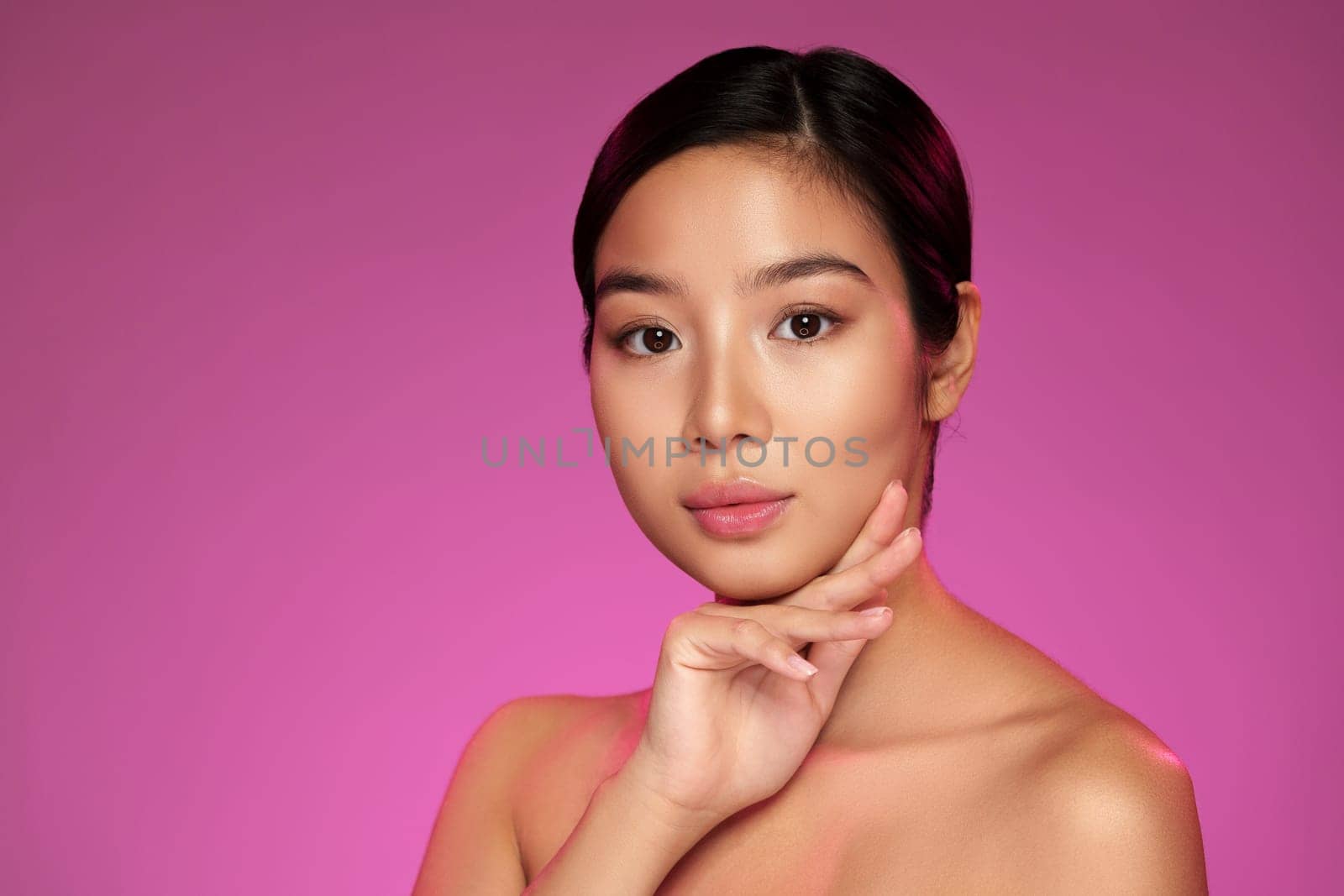 A confident woman poses gracefully with her hand touching her neck, framed by a bright pink backdrop. The expression captures elegance and poise, highlighting her features beautifully.