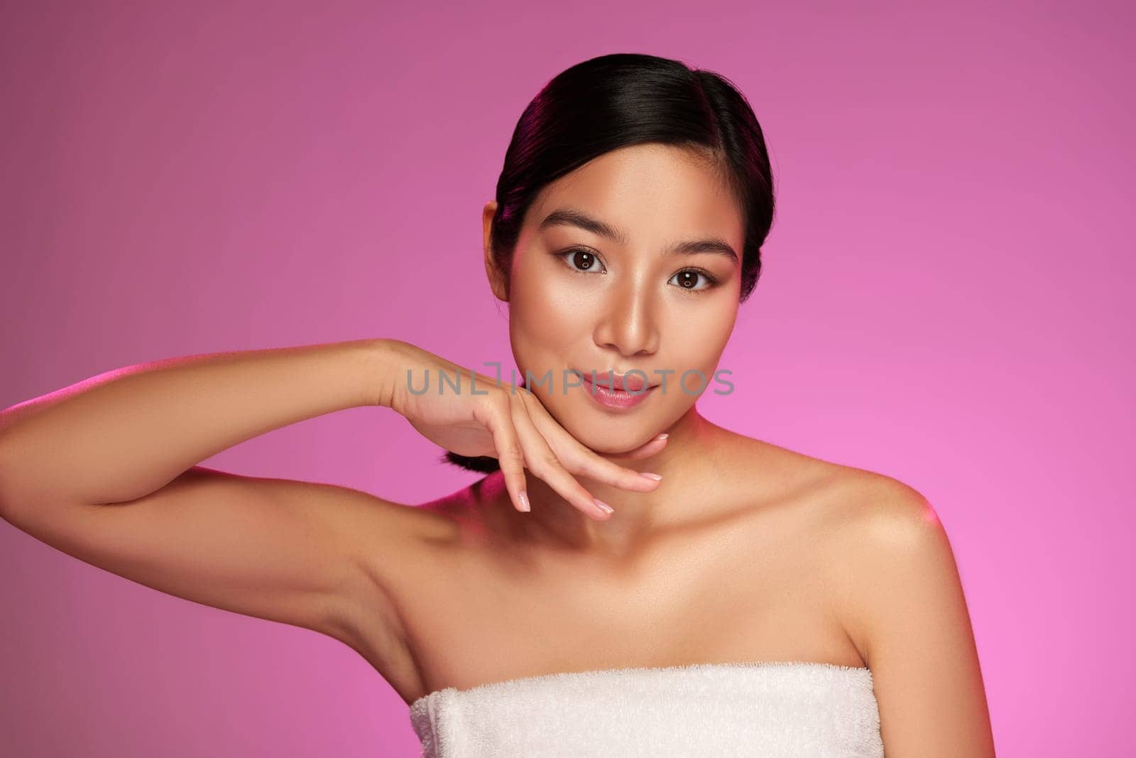A model showcases a stylish pose with one hand resting elegantly on her chin, illuminated by a soft pink light. Her expression is poised and confident, highlighting beauty and grace.