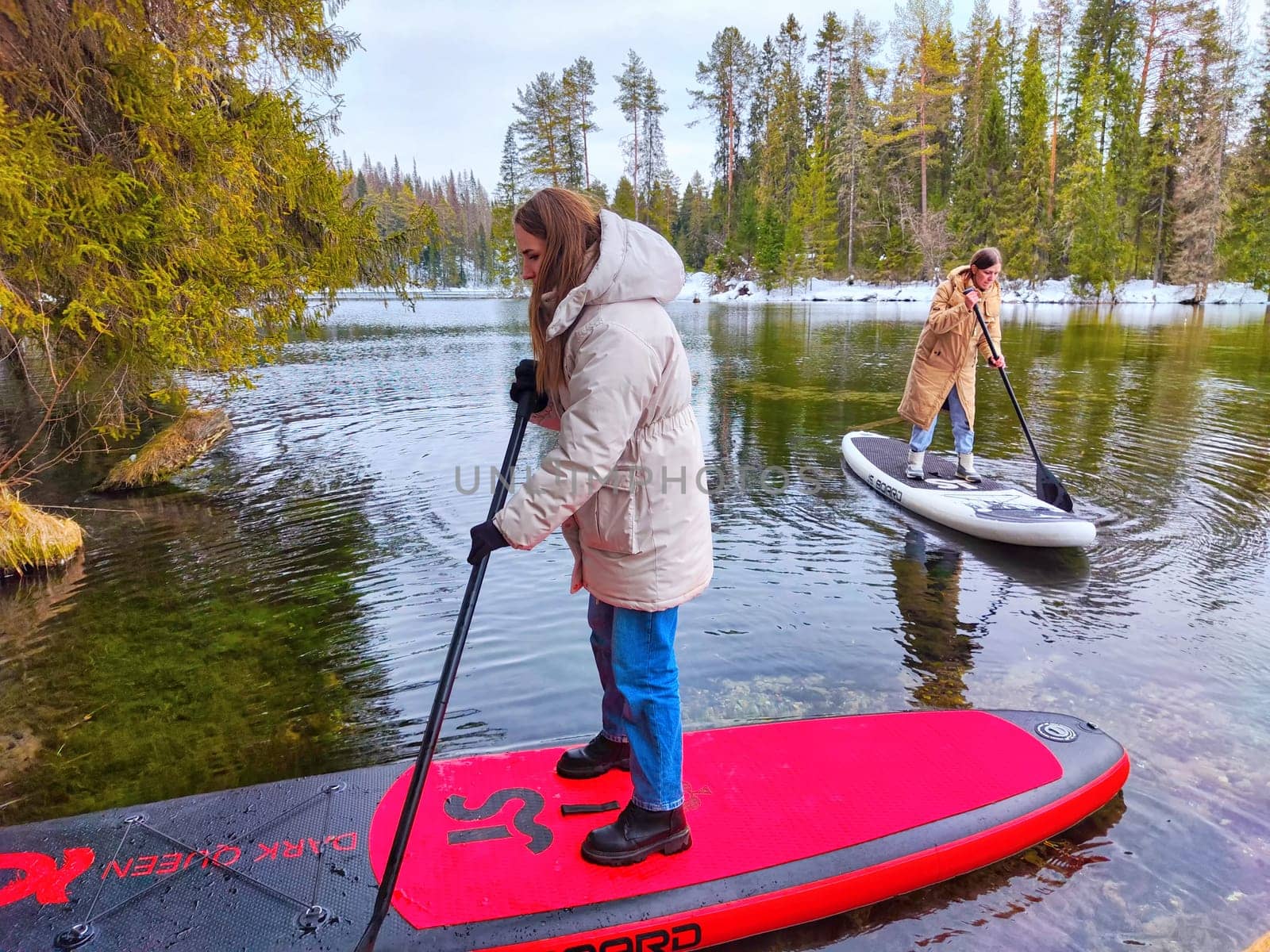 Kirov, Russia - March 14, 2024: Two people are paddle boarding on a serene lake with vibrant greenery and clear blue skies, enjoying a refreshing outdoor experience.