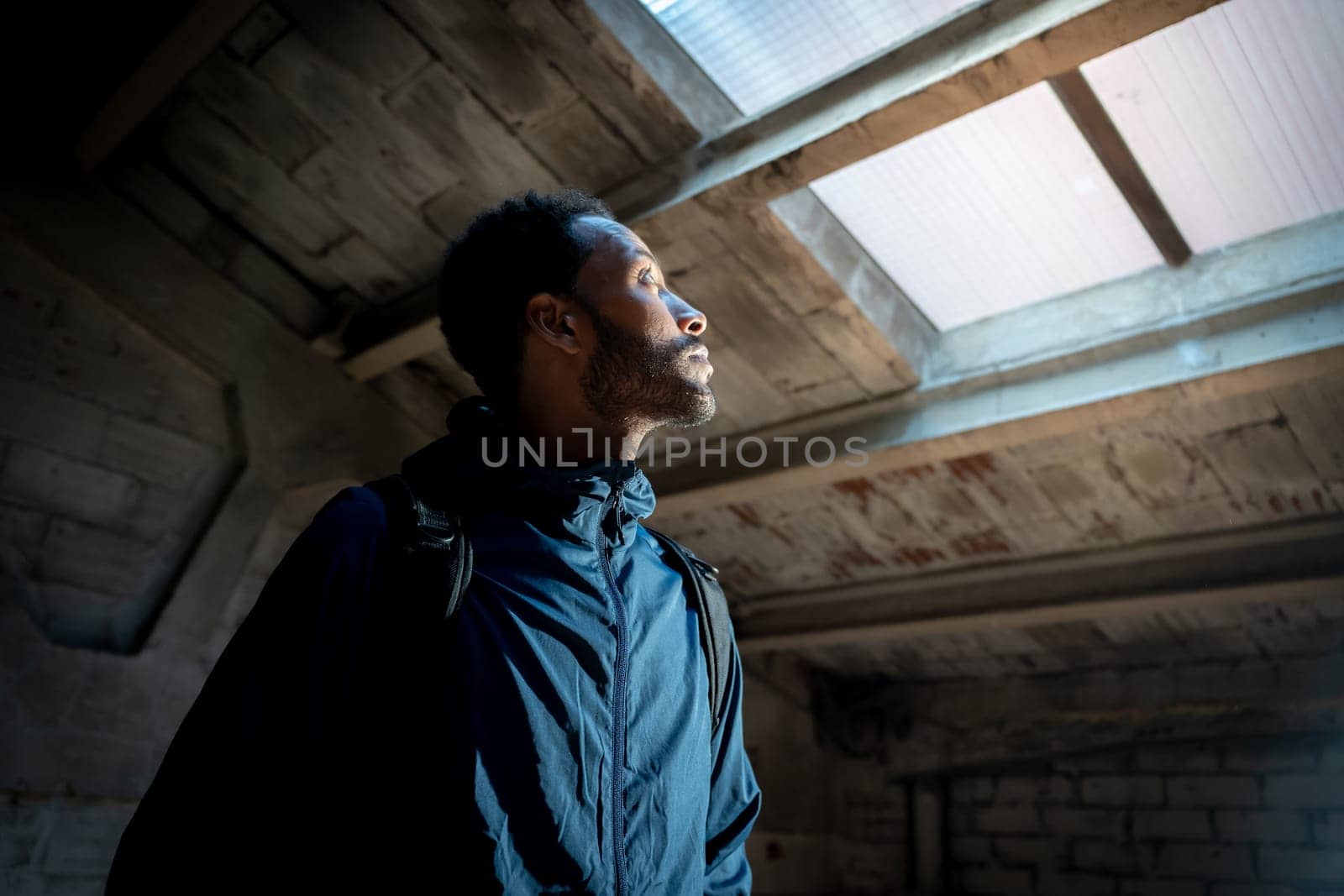 A contemplative man thoughtfully gazes upwards towards a skylight window situated within a dimly lit, quiet space